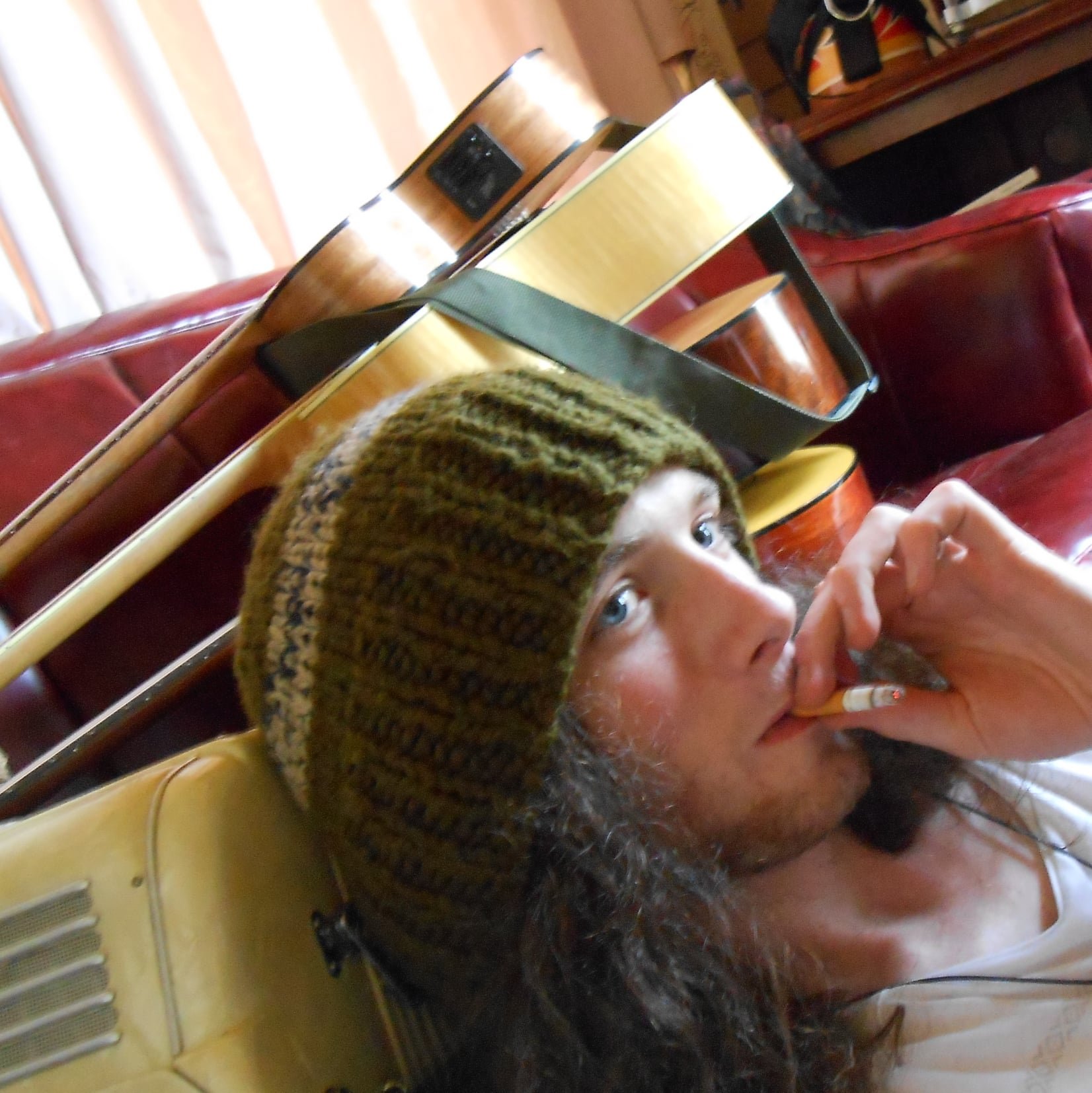 A young man with long hair and a knitted beanie hat is sitting on a yellow chair, holding a cigarette in his mouth and smoking while looking at the camera. A guitar rests on the chair behind him, and red leather couches are visible in the background.
