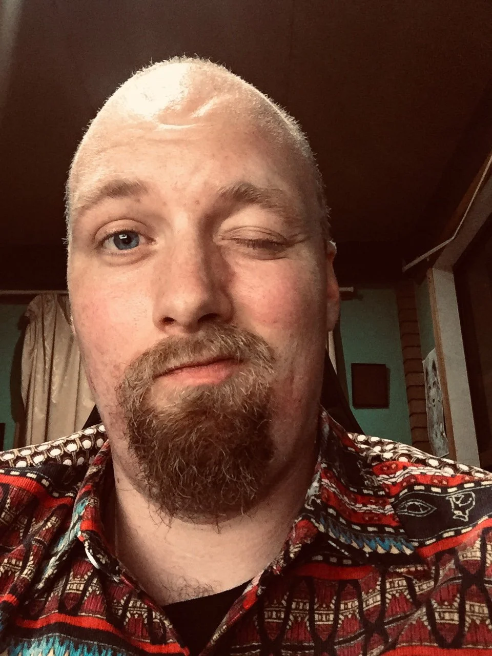 A man with a light beard and mustache, one blue eye visible, the other eye partially closed. Wearing a colorful patterned shirt, in a room with wooden ceiling and walls, and a curtain in the background.