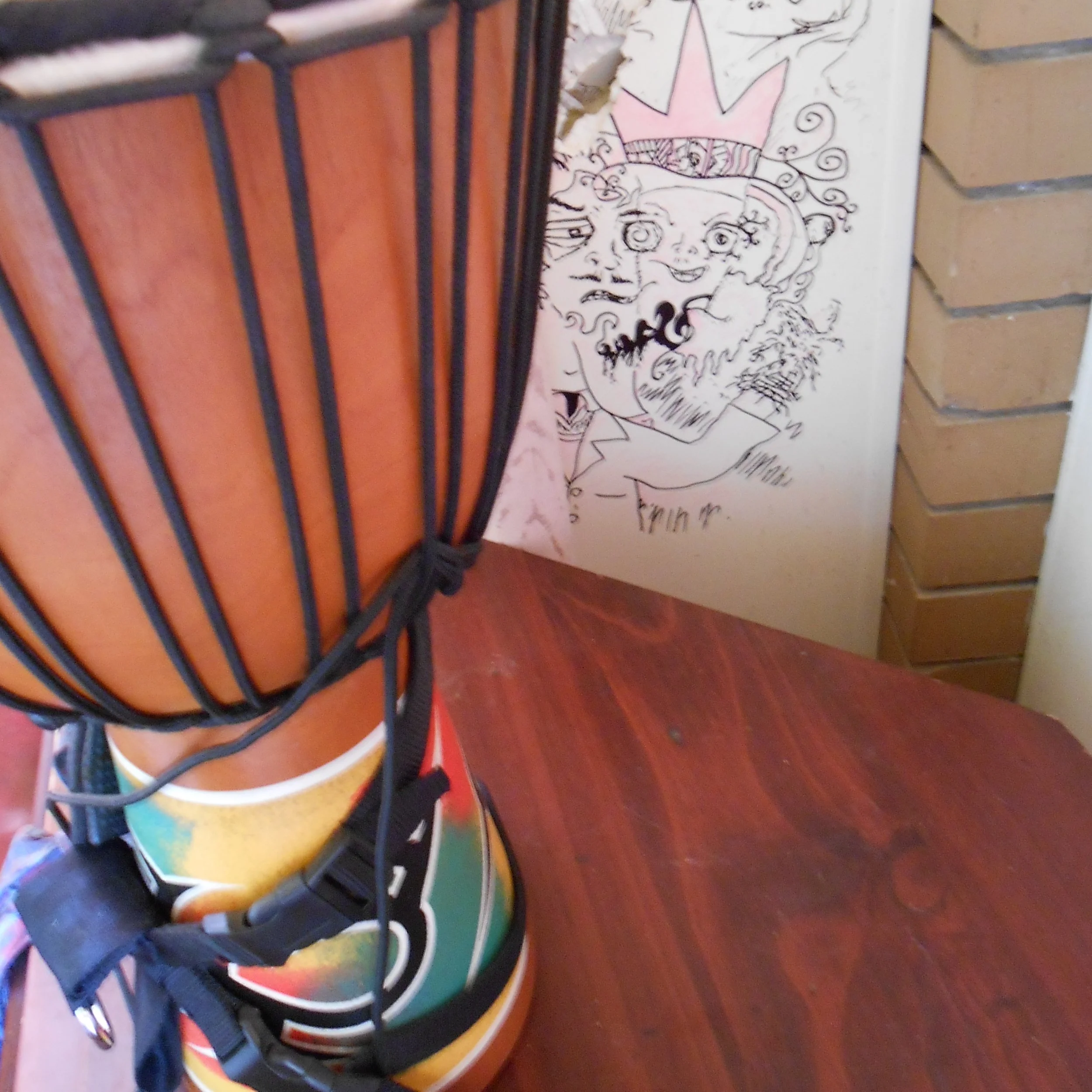 A colorful lamp with a striped orange and black shade on a wooden table, with a drawing of a smiling, crowned character on the wall in the background.