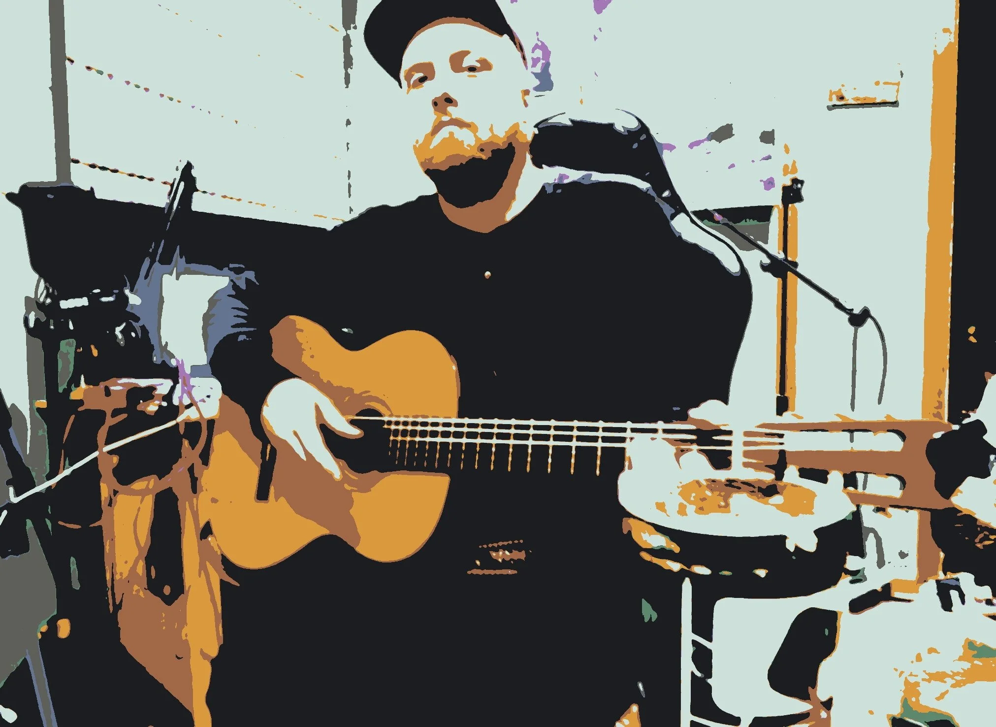 A person playing an acoustic guitar in a room with musical equipment around, microphone in front, and a light-colored wall with some items hanging.