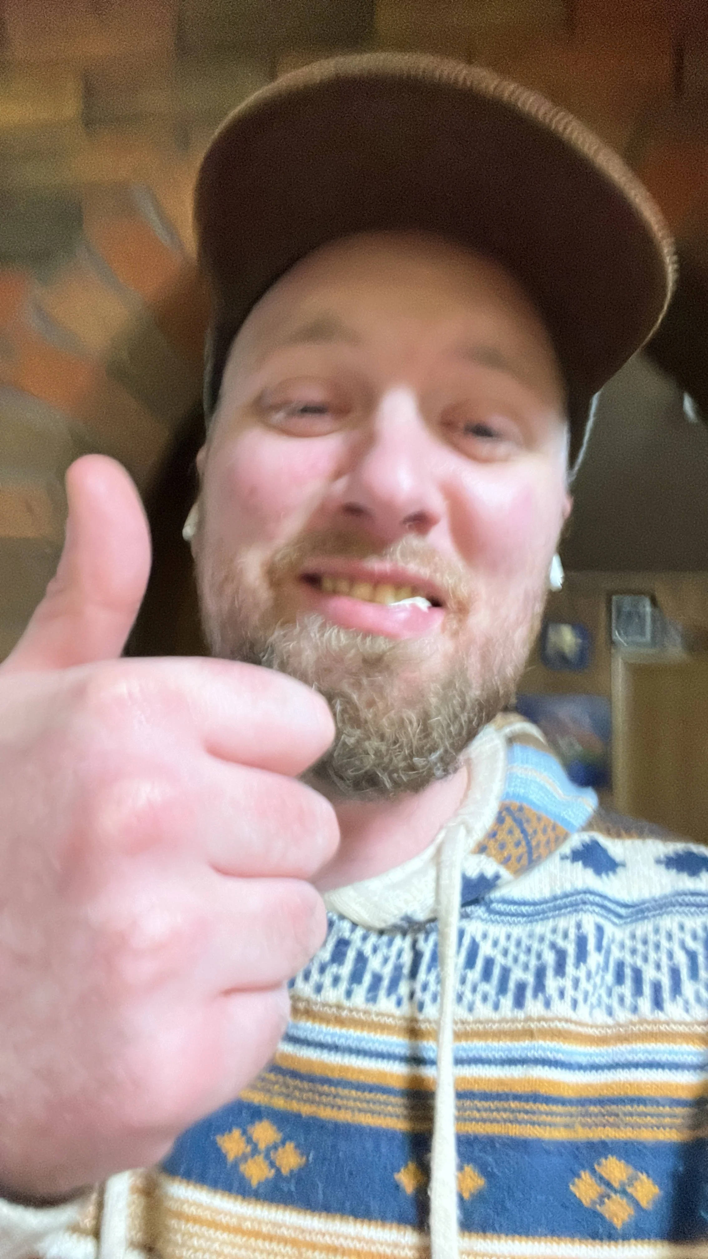 A man with a beard wearing a patterned sweater and a baseball cap, giving a thumbs-up and smiling indoors.