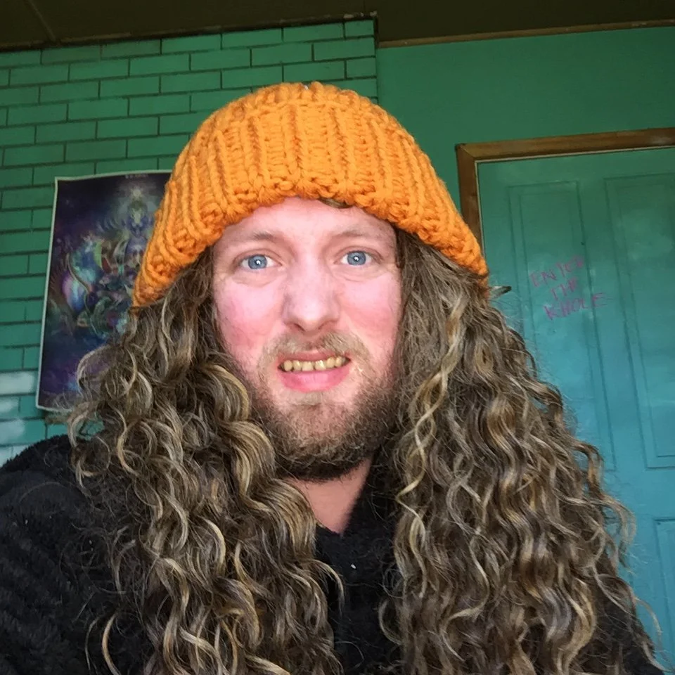 A man with long, curly hair wearing a bright orange knit beanie and a black jacket. He has a beard and is smiling with his teeth showing. The background features a green brick wall, a framed artwork, and a blue door with pink writing.