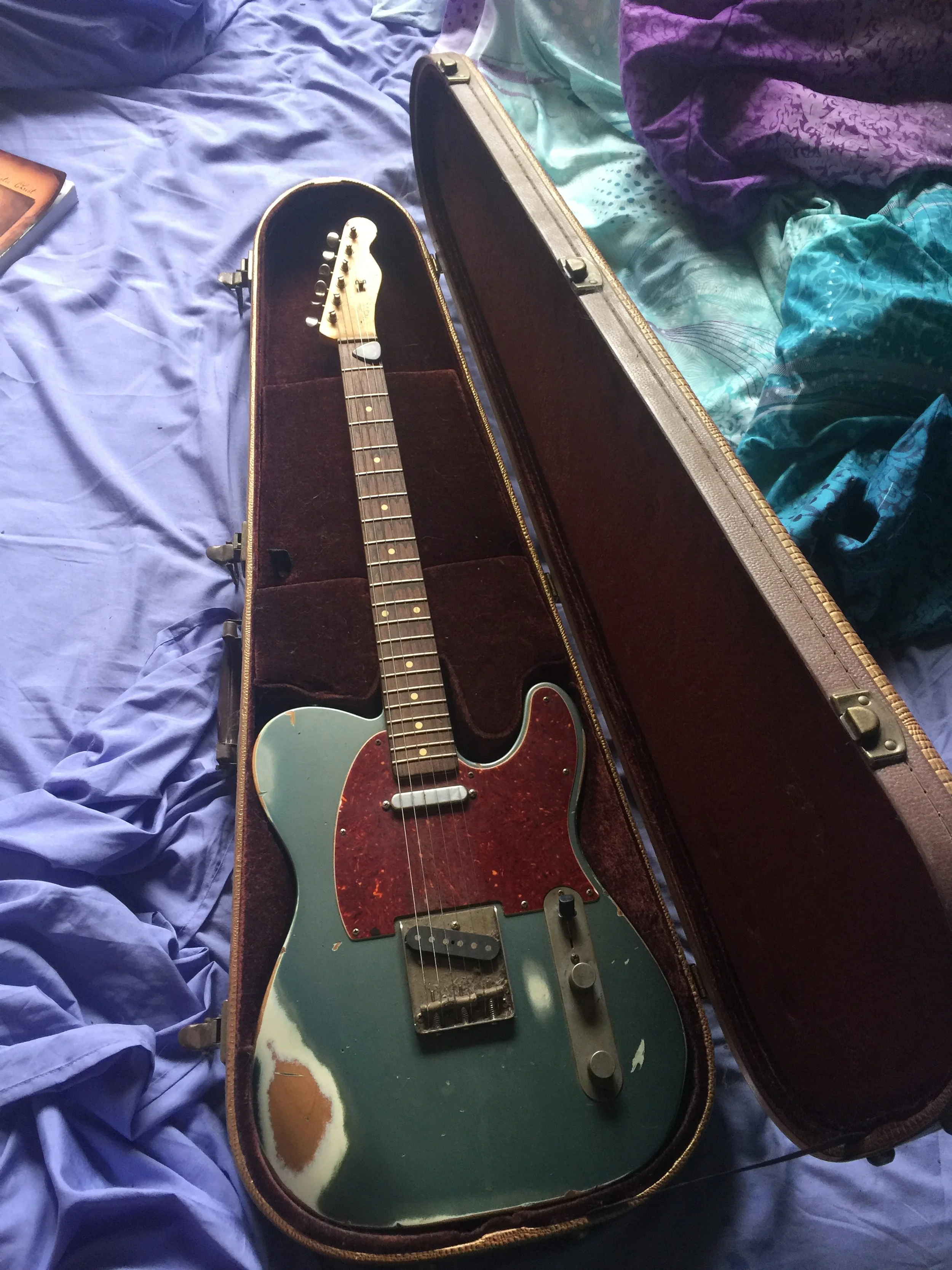 An electric guitar with a worn green body and a red tortoiseshell pickguard in an open vintage hard case, resting on a bed with purple, teal, and patterned sheets.