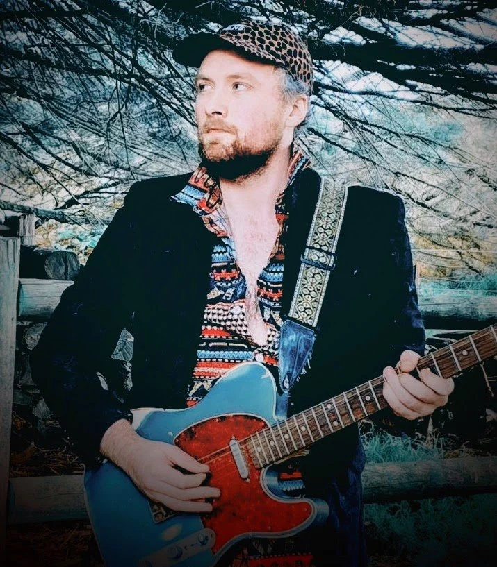 A man with a beard wearing a leopard print cap, a patterned shirt, and a black jacket holding an electric guitar outdoors with trees and wooden logs in the background.