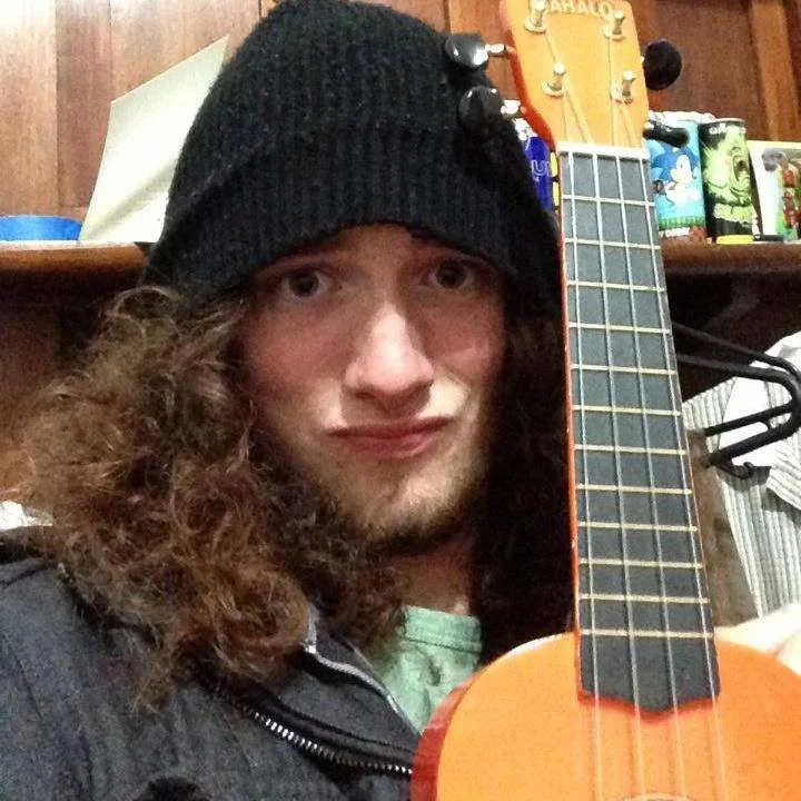 A young man with long curly hair, wearing a black beanie and black jacket, holding an acoustic guitar indoors with a wooden wall and various household items in the background.