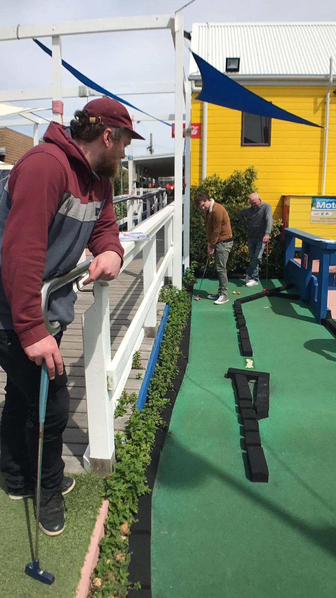 Three men playing mini golf outdoors on a sunny day. One man in the foreground is leaning on a white railing, smiling, while two others in the background are putting on a green artificial turf.