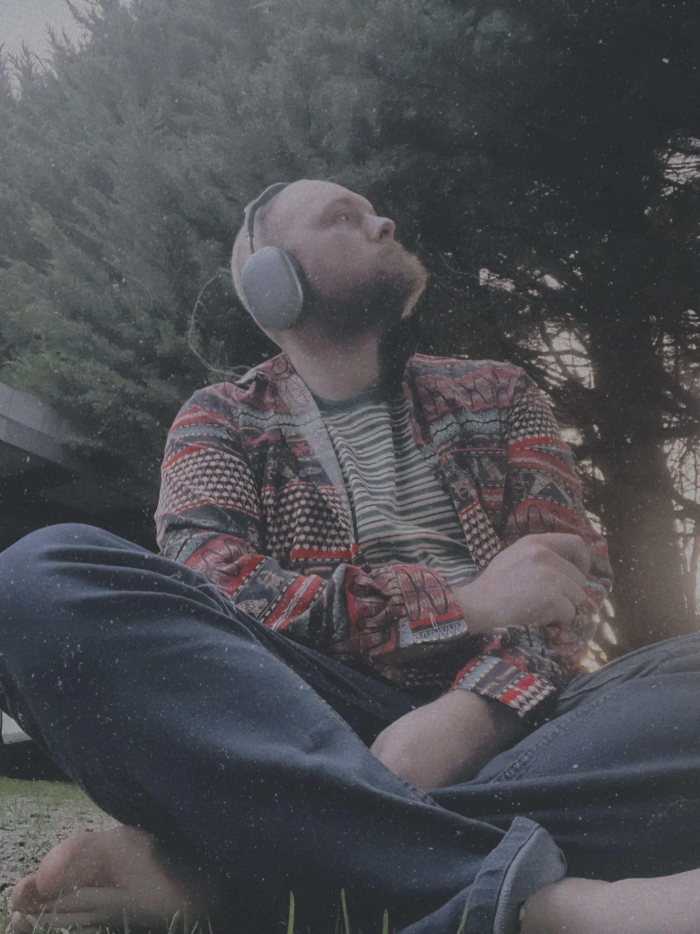 A man with a beard and headphones relaxing outdoors, sitting cross-legged on the grass, wearing a patterned jacket and striped shirt, with trees in the background.