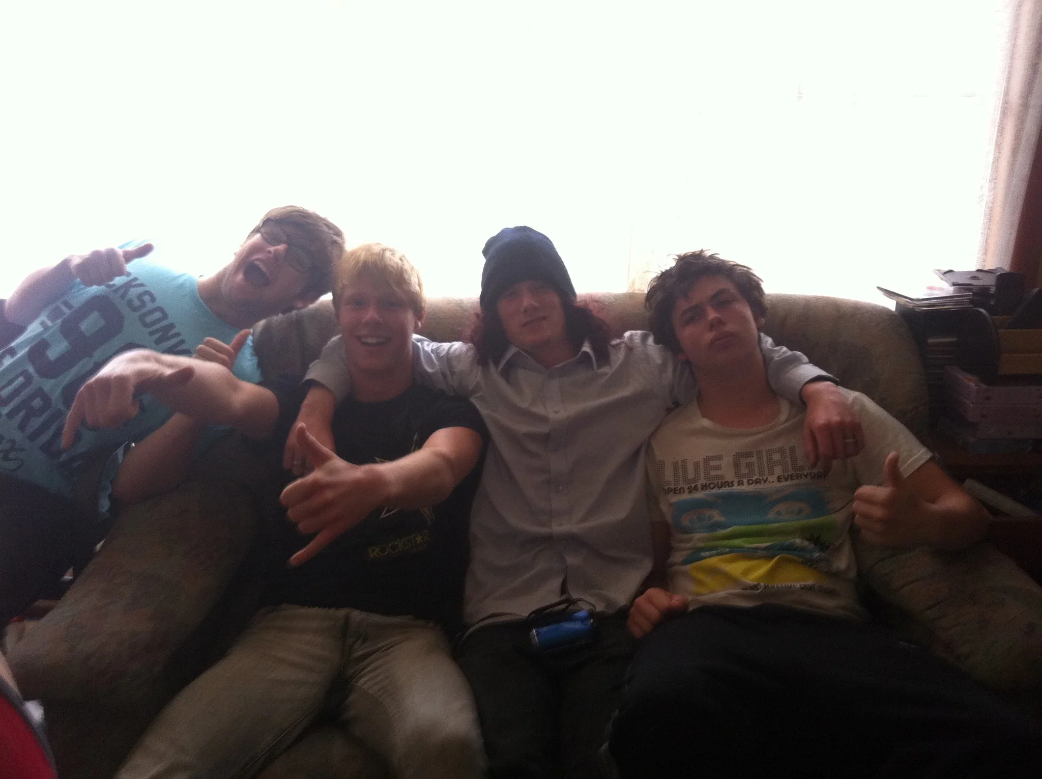 Five young men sitting on a couch, smiling and making various hand gestures, with a bright window behind them and some stacked items on the side.