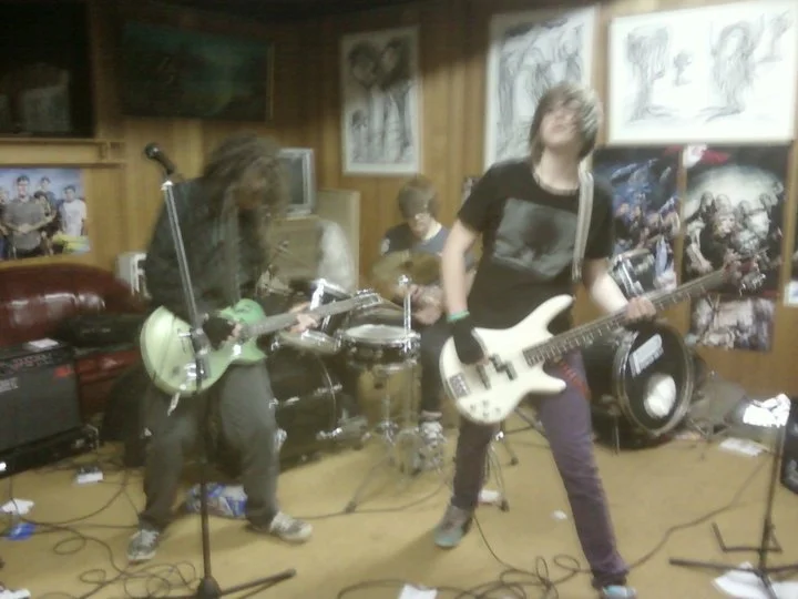 Three young musicians playing electric guitars and drums in a wood-paneled room with posters and artwork on the walls.