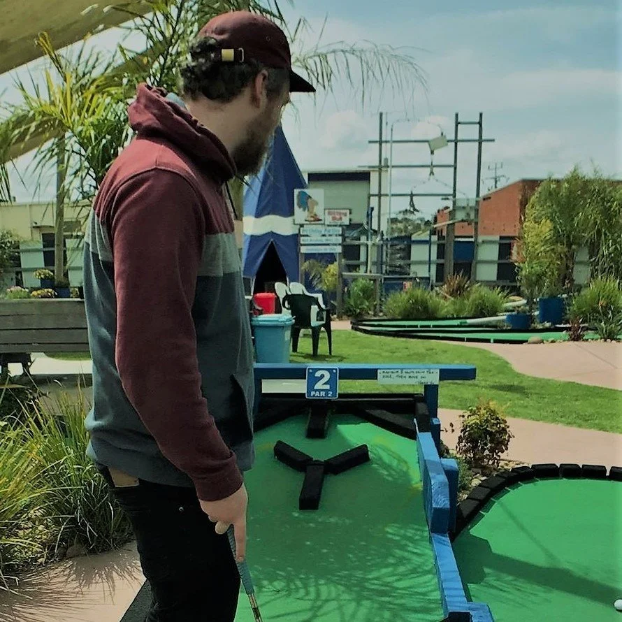 Man playing mini golf on a putting green, holding a golf club, and aiming at a hole marked with a blue sign that reads '2 PAR 2'.
