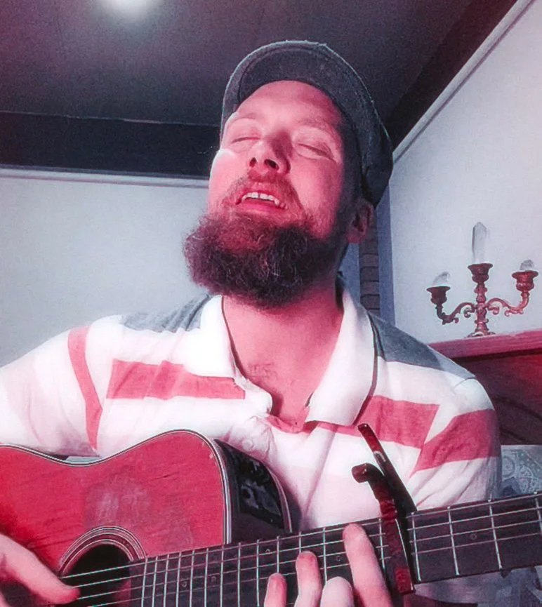 A man with a beard and mustache wearing a cap and a striped polo shirt, playing an acoustic guitar with his eyes closed and a relaxed expression.