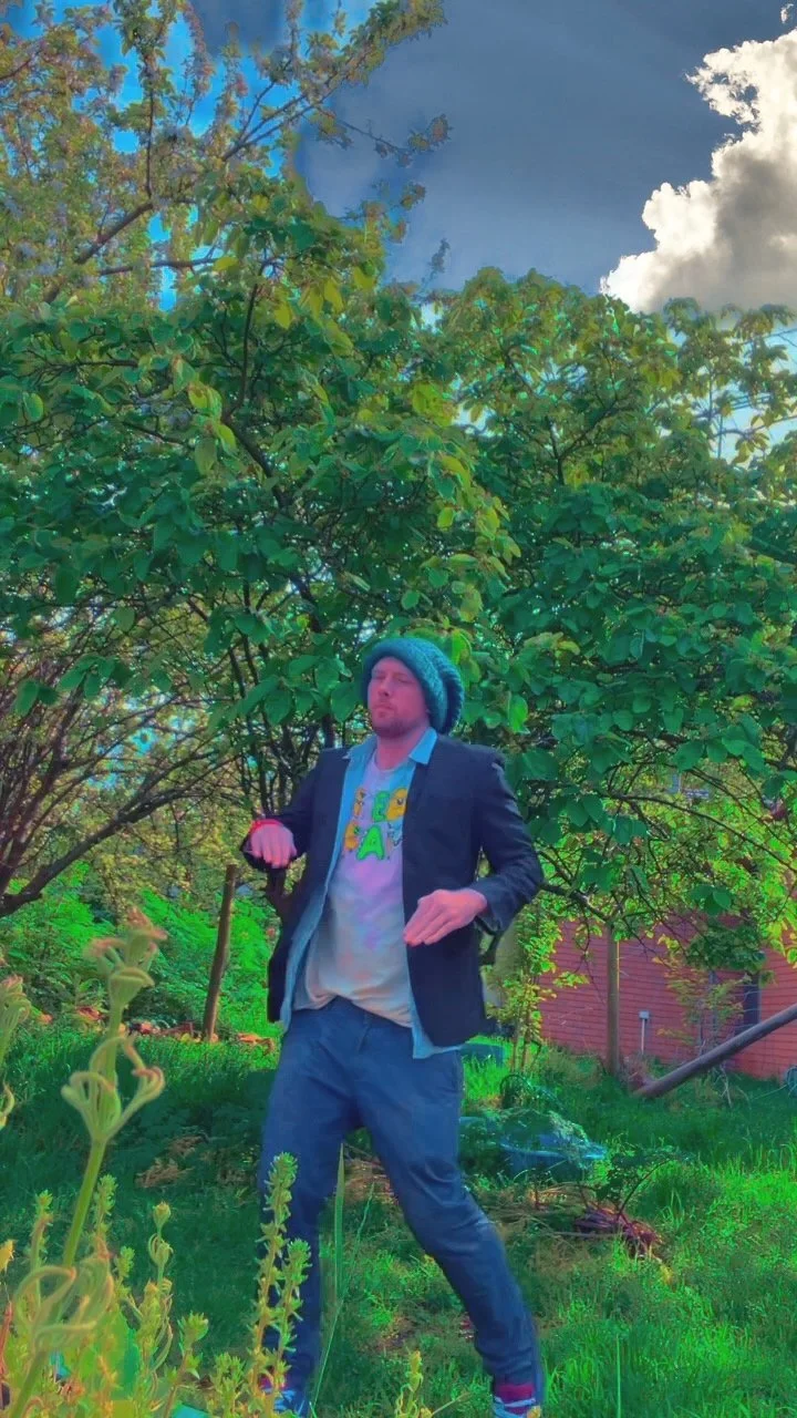 A man stands outdoors in front of green trees, wearing a dark blazer, a colorful T-shirt, blue jeans, a beanie hat, and gloves. He appears to be dancing or in a relaxed pose with eyes closed.