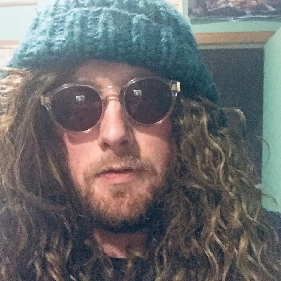 A person with long, curly hair wearing dark sunglasses and a teal knit hat.
