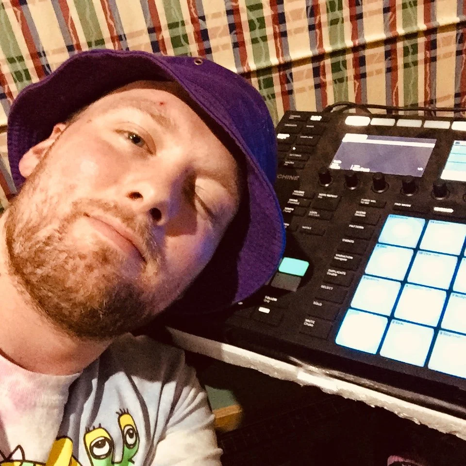 A man wearing a purple bucket hat and a graphic t-shirt with cartoon eyes, taking a selfie next to a music production controller with blue-lit pads and various knobs.