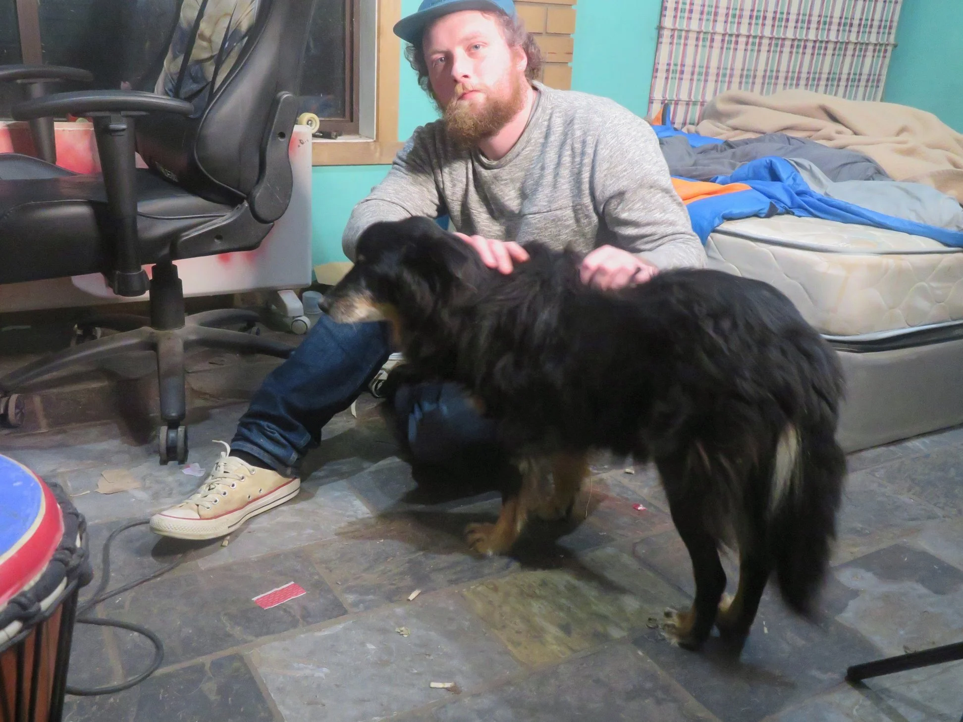 A man with a beard and wearing a cap, grey sweater, and jeans, sits on the floor petting a large black dog inside a cluttered bedroom. The room has an unmade bed with blankets, a window with a wooden frame, an office chair, and a tiled floor.