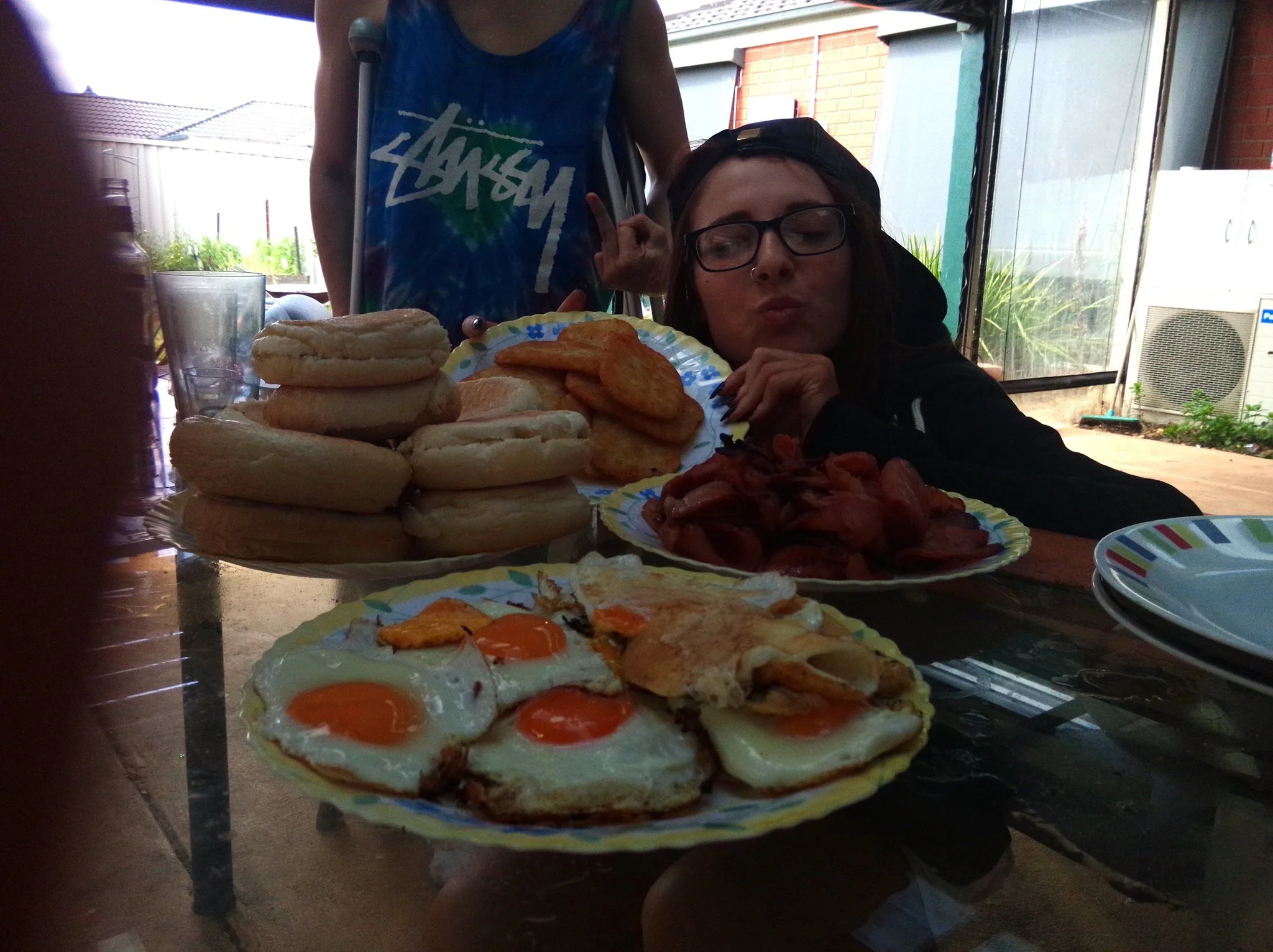 A woman with glasses and a black cap sitting at a table with breakfast foods including fried eggs, toast, bacon, and potato wedges, with a woman in a blue tie-dye shirt and glasses behind her making a peace sign.