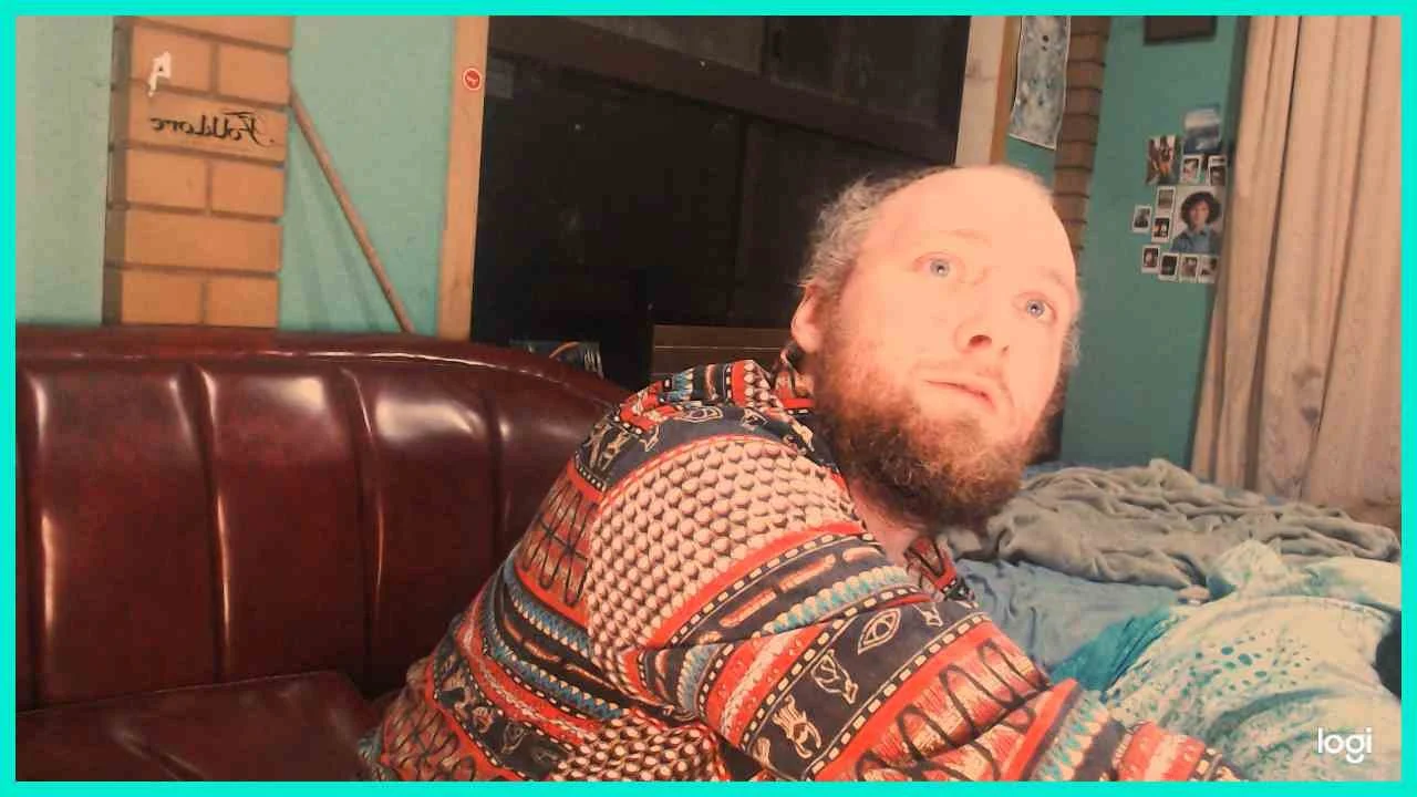 A man with a beard and receding hairline looking surprised or confused, sitting on a leather couch in a room with teal walls, a window behind him, and photos on the wall.