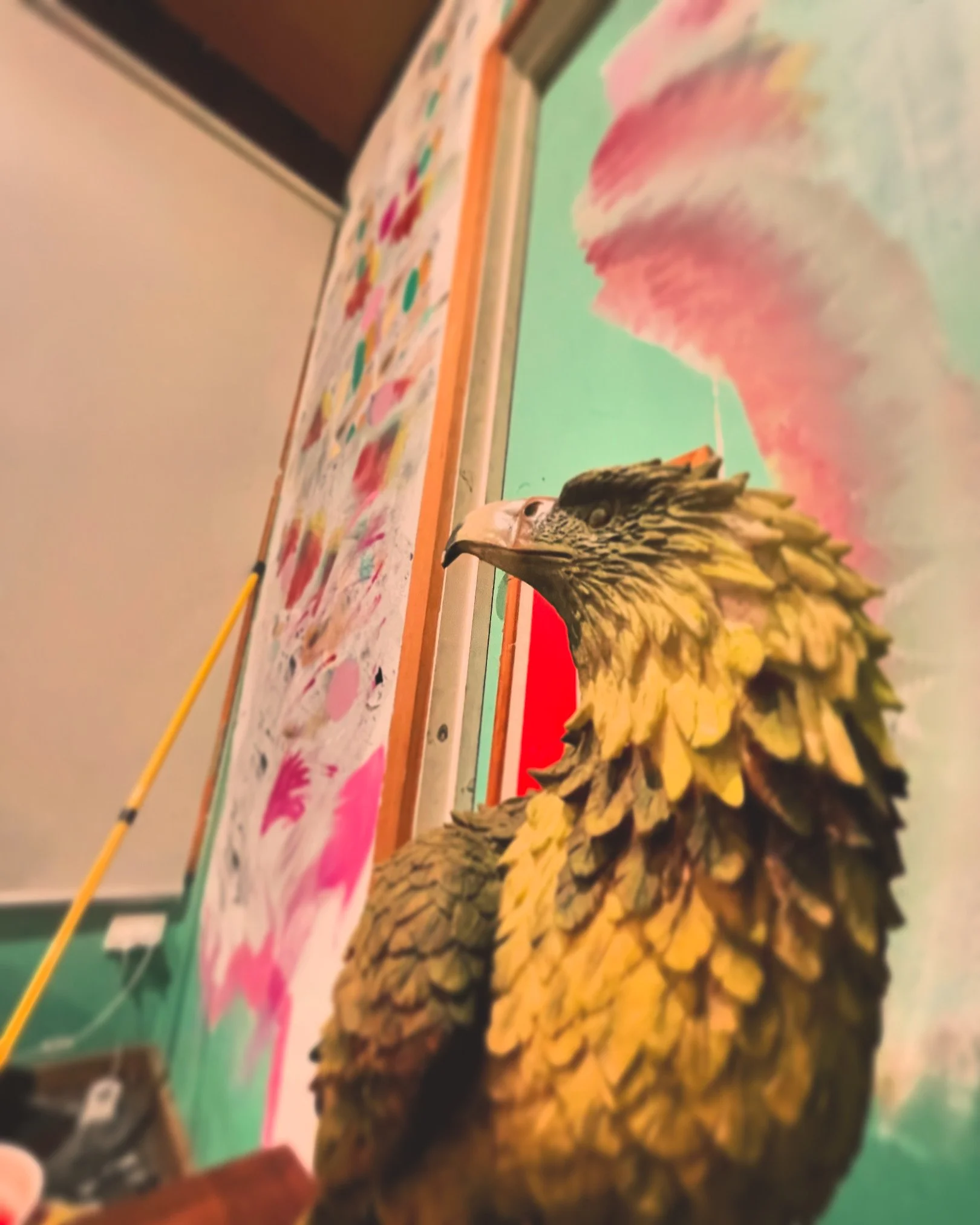 Close-up of a three-dimensional eagle sculpture with detailed feathers. In the background, a colorful wall with abstract pink, green, and white graphics and a whiteboard with various marks and a long yellow pointer leaning against it.