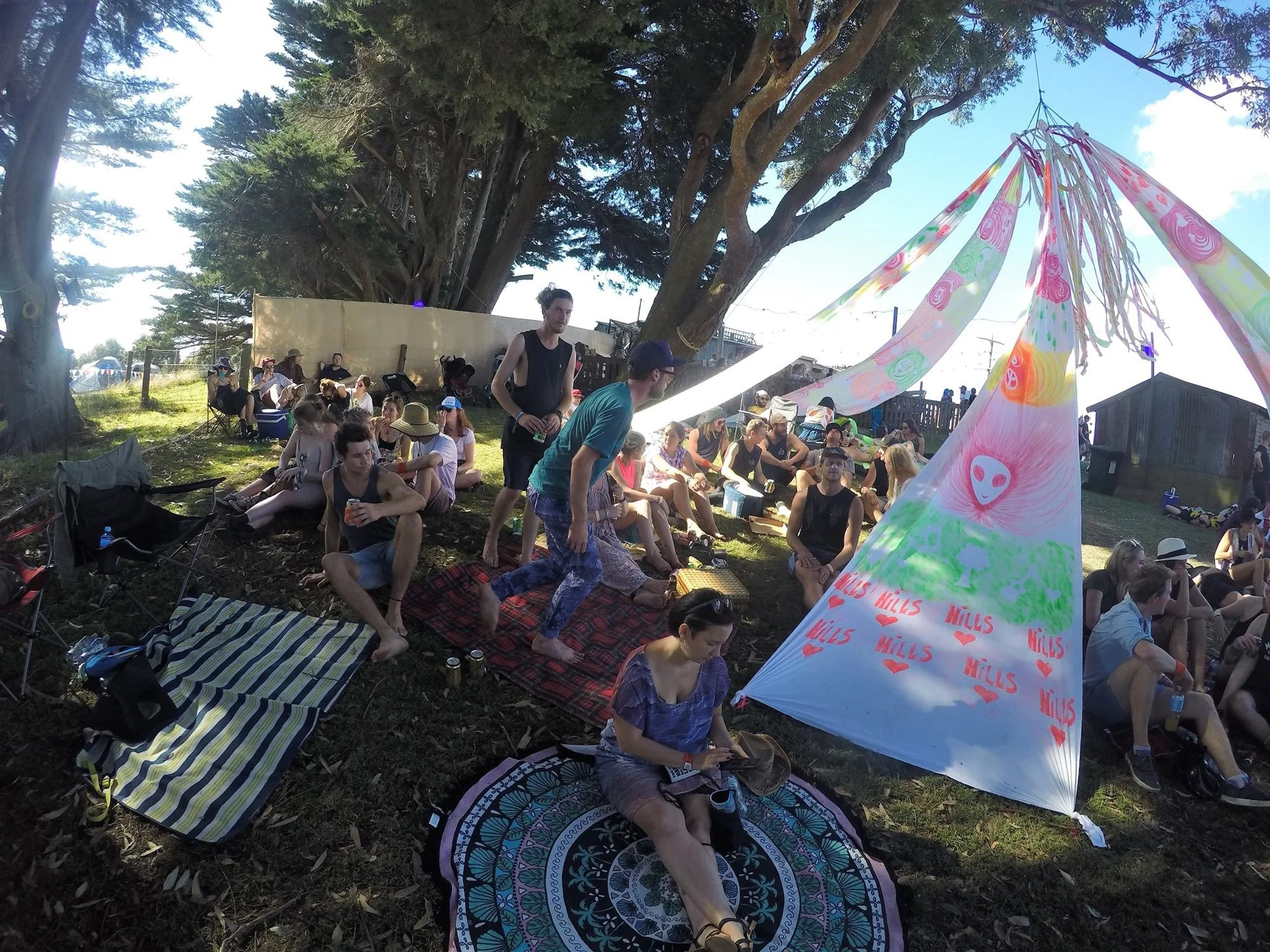 People sitting on the grass under trees at an outdoor event, with a colorful fabric canopy decorated with drawings and the word 'HILLS'. Some people are socializing and drinking, and there are tents and structures in the background.