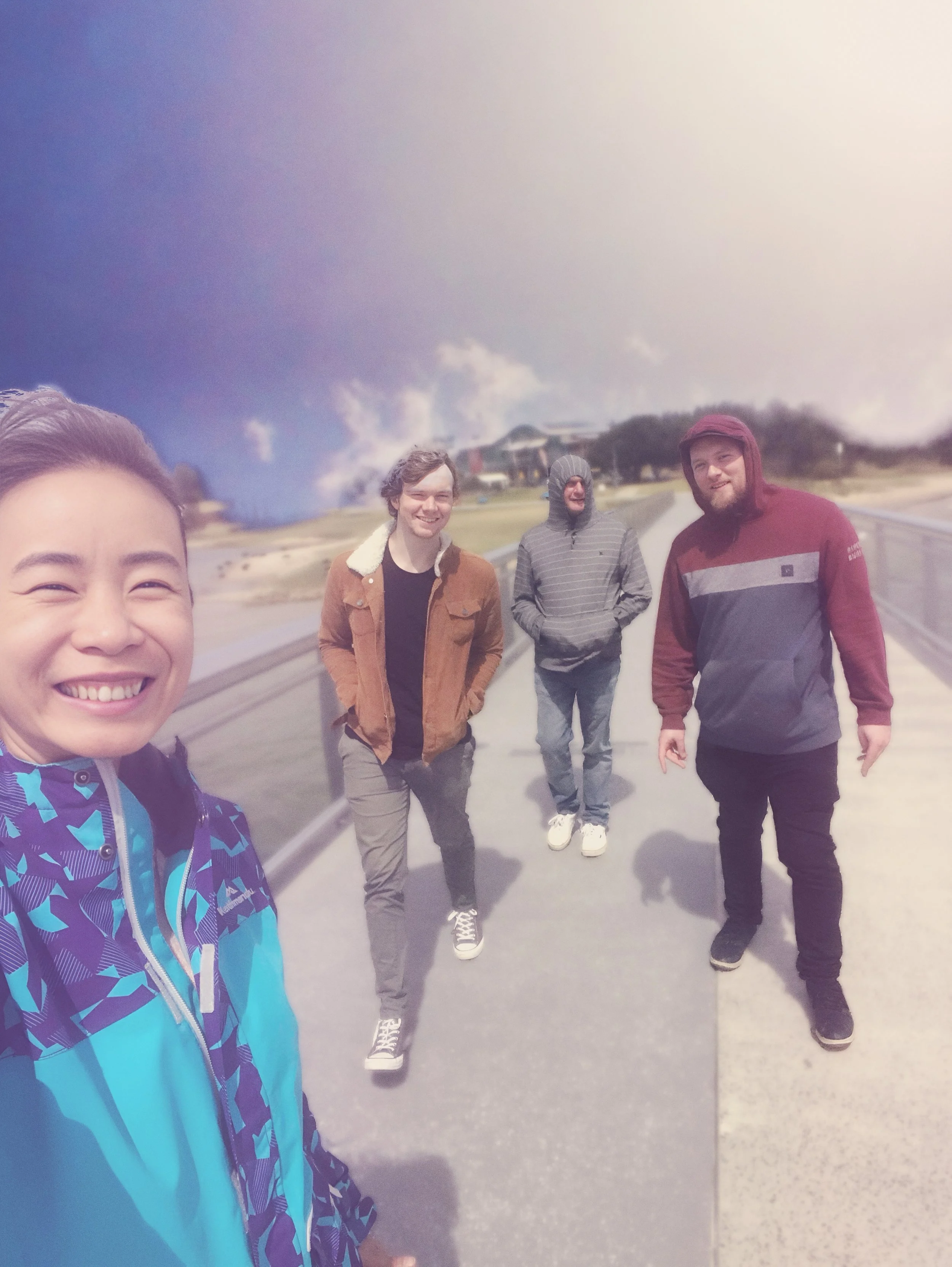 Four people standing on a bridge outdoors, with a cloudy sky in the background. One person is taking a selfie, and the others are smiling.