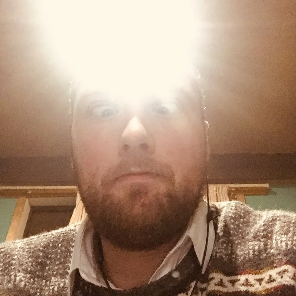 A man with a beard and mustache looking at the camera with wide eyes. Bright light from overhead creates a glare on his face. He is wearing a patterned sweater over a collared shirt, in a room with wooden paneling and a door in the background.