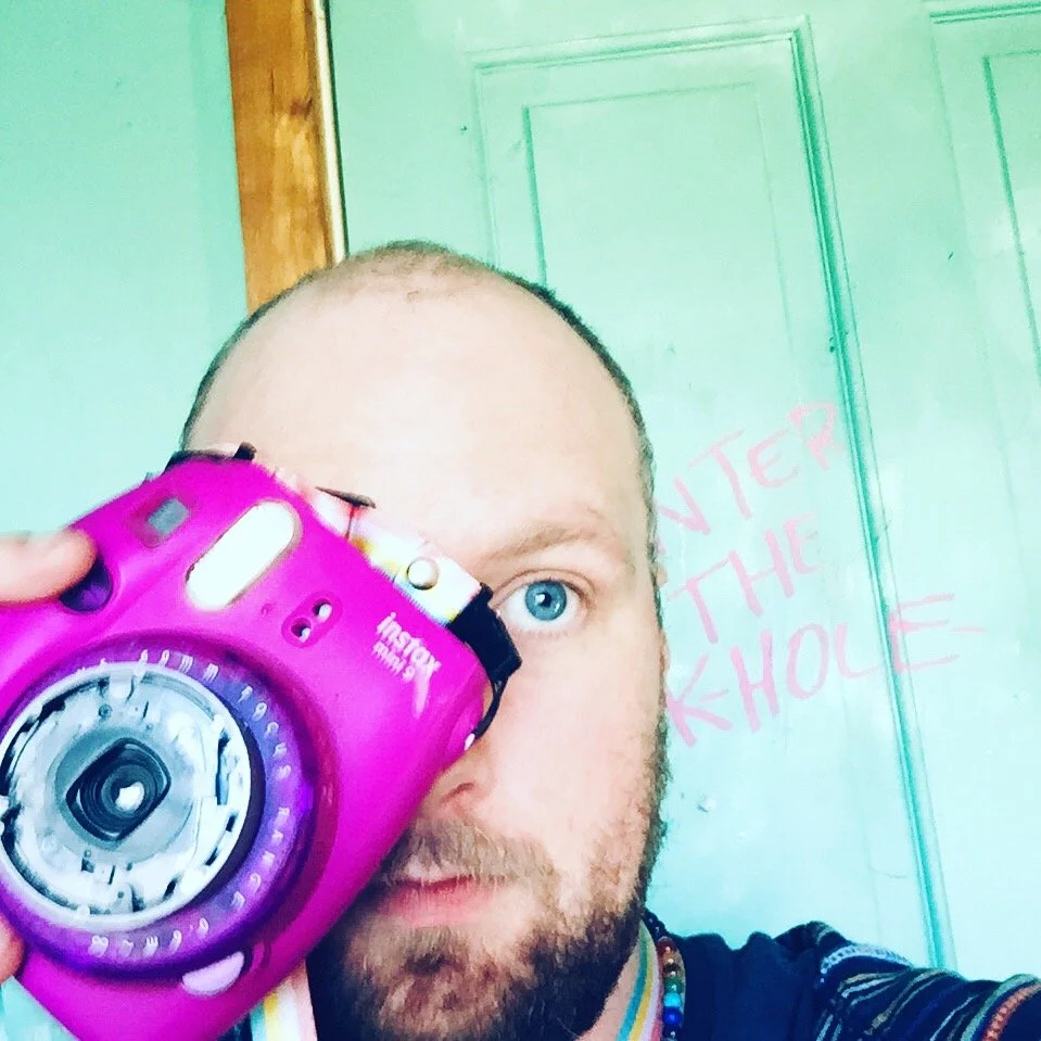 A man with a beard and blue eyes holding a pink instant camera partially covering his face, standing in front of a light green door with pink text that reads 'ENTER THE SCHOOL'.