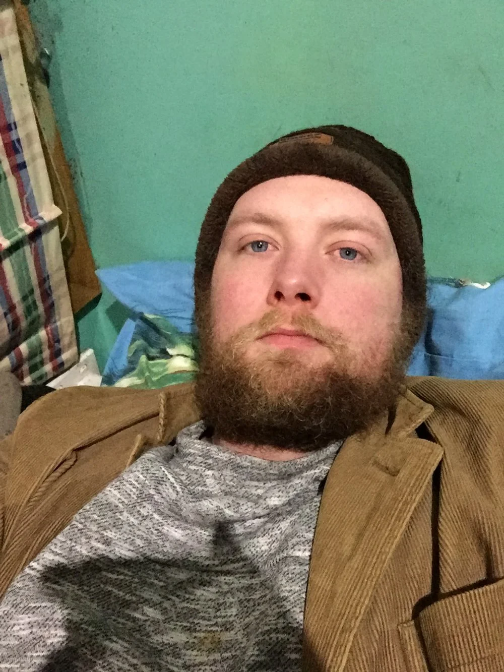 A man with a beard and blue eyes lying on a bed with green walls. He is wearing a brown jacket, a gray shirt, and a black beanie. There are patterned curtains and a backpack visible in the background.