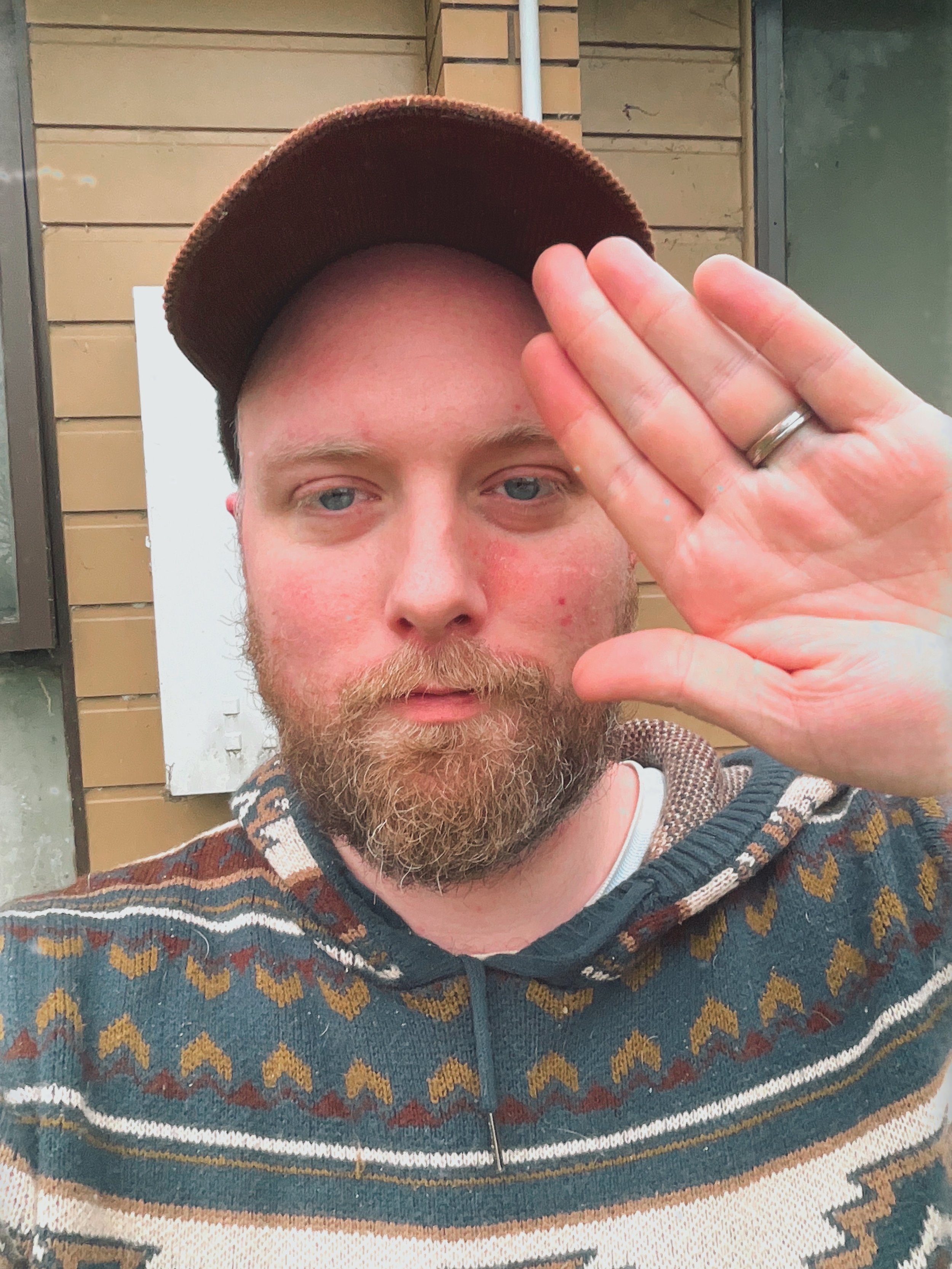 A man with a beard and wearing a patterned sweater and a backward baseball cap, holding up his hand with a wedding ring visible, standing in front of a building with wood siding.