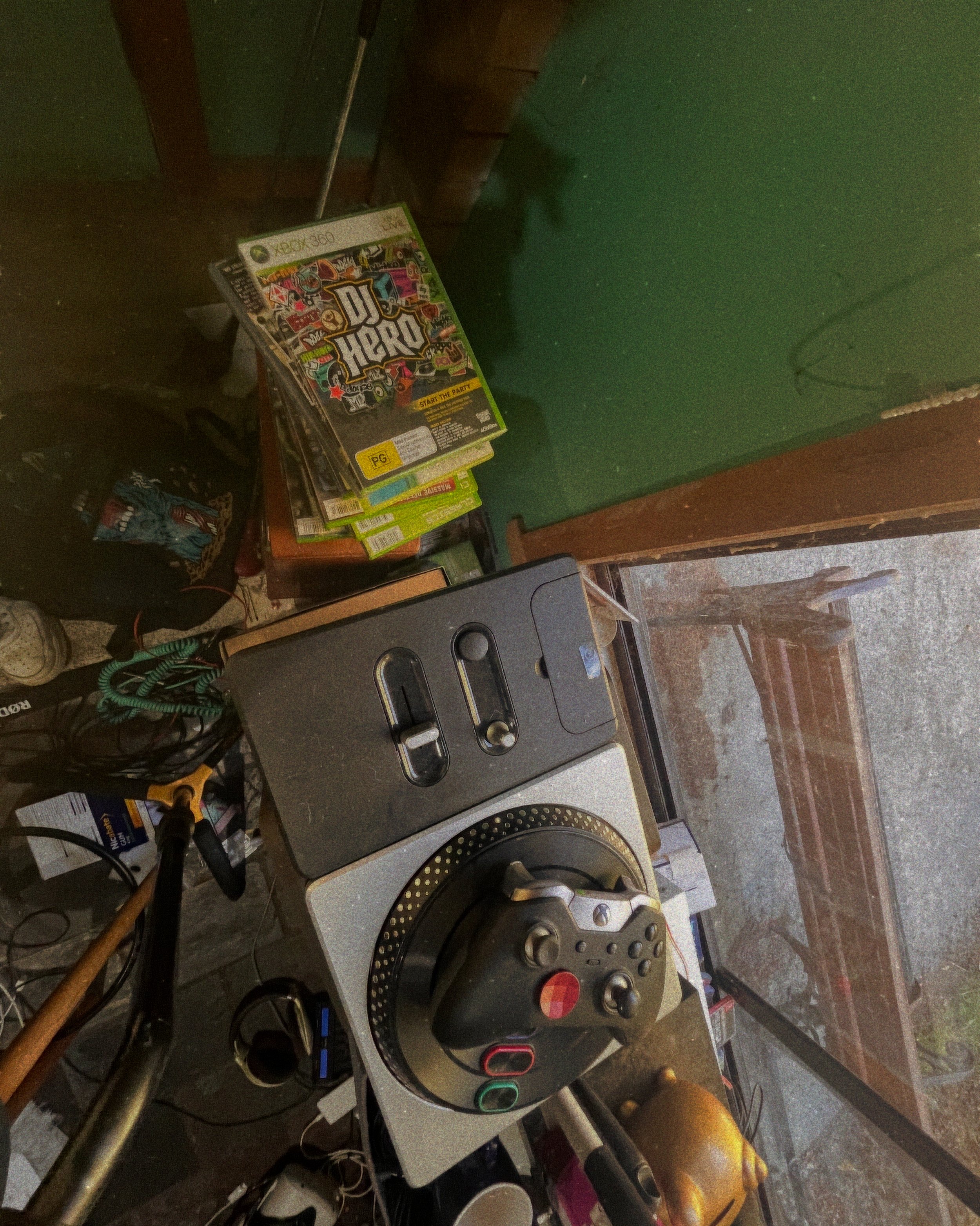A turntable with a game controller placed on it, next to a small pile of video games, and other miscellaneous items on a cluttered table in a room with a green wall and a window showing an outdoor bench.
