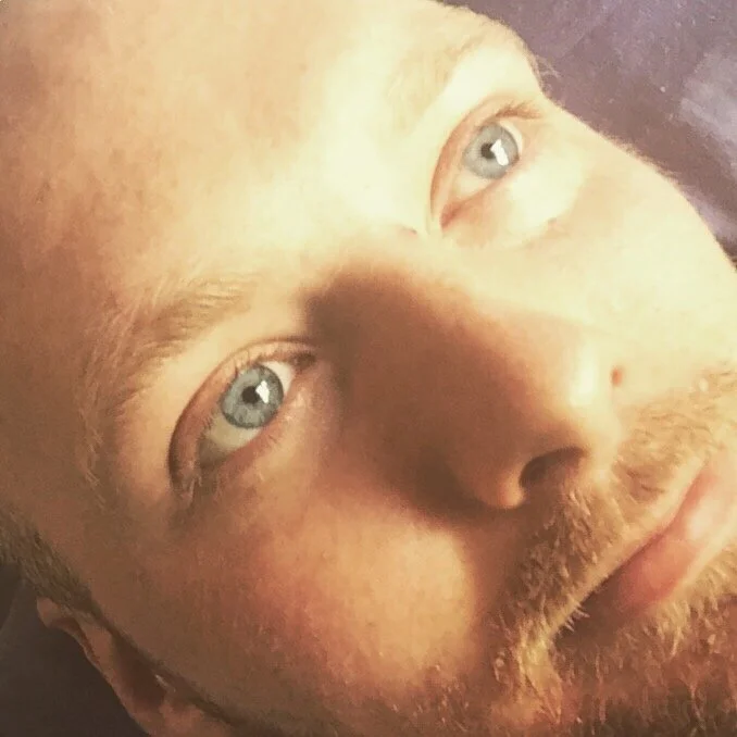 Close-up of a man's face with light skin, blue eyes, and a beard, lying down and looking upwards.
