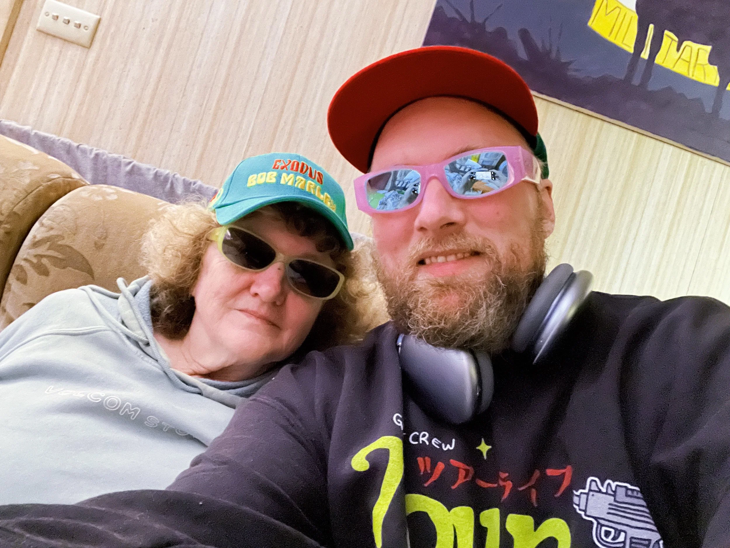 A man and a woman taking a selfie indoors. They are wearing sunglasses, and the man is also wearing a red cap and headphones around his neck. The woman is wearing a colorful cap that says 'EXODUS' and 'BOB MARLEY' and a hoodie. There is a poster on t