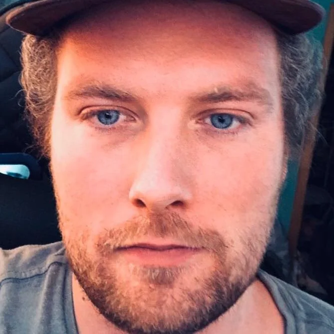 Close-up of a young man with blue eyes, light brown hair, a beard, wearing a dark cap and a gray shirt.