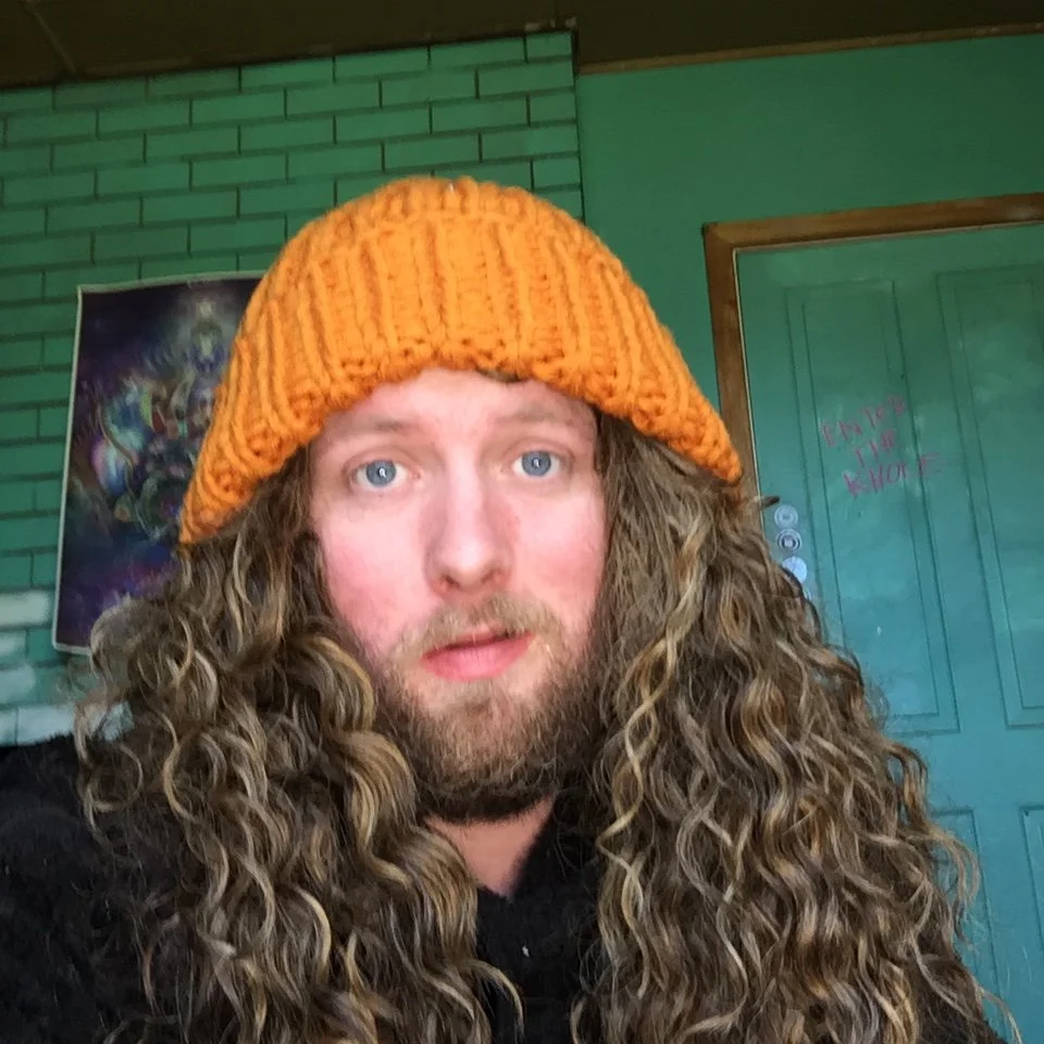 A person with long curly hair and a beard wearing an orange knit hat in a room with green brick walls and a teal door.