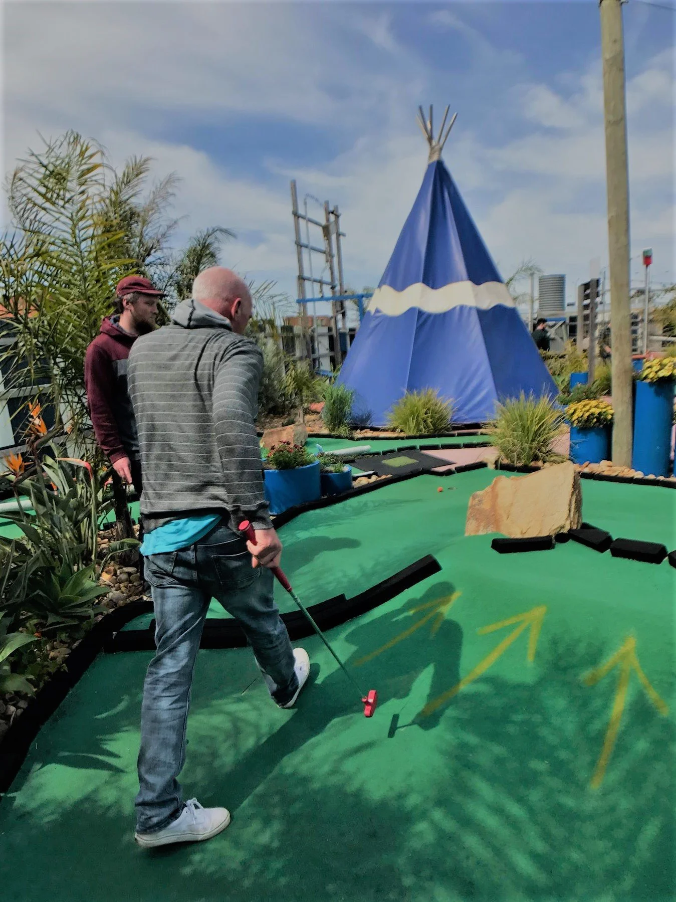 People playing mini golf on an outdoor course with a teeing area, plants, and a blue teepee structure in the background under a partly cloudy sky.