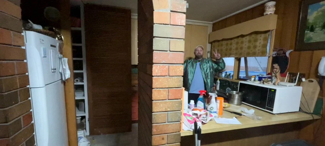 A man with a beard making a peace sign behind a kitchen counter, with a brick column in the foreground, in a cozy kitchen with wooden walls, a microwave, and various kitchen items.