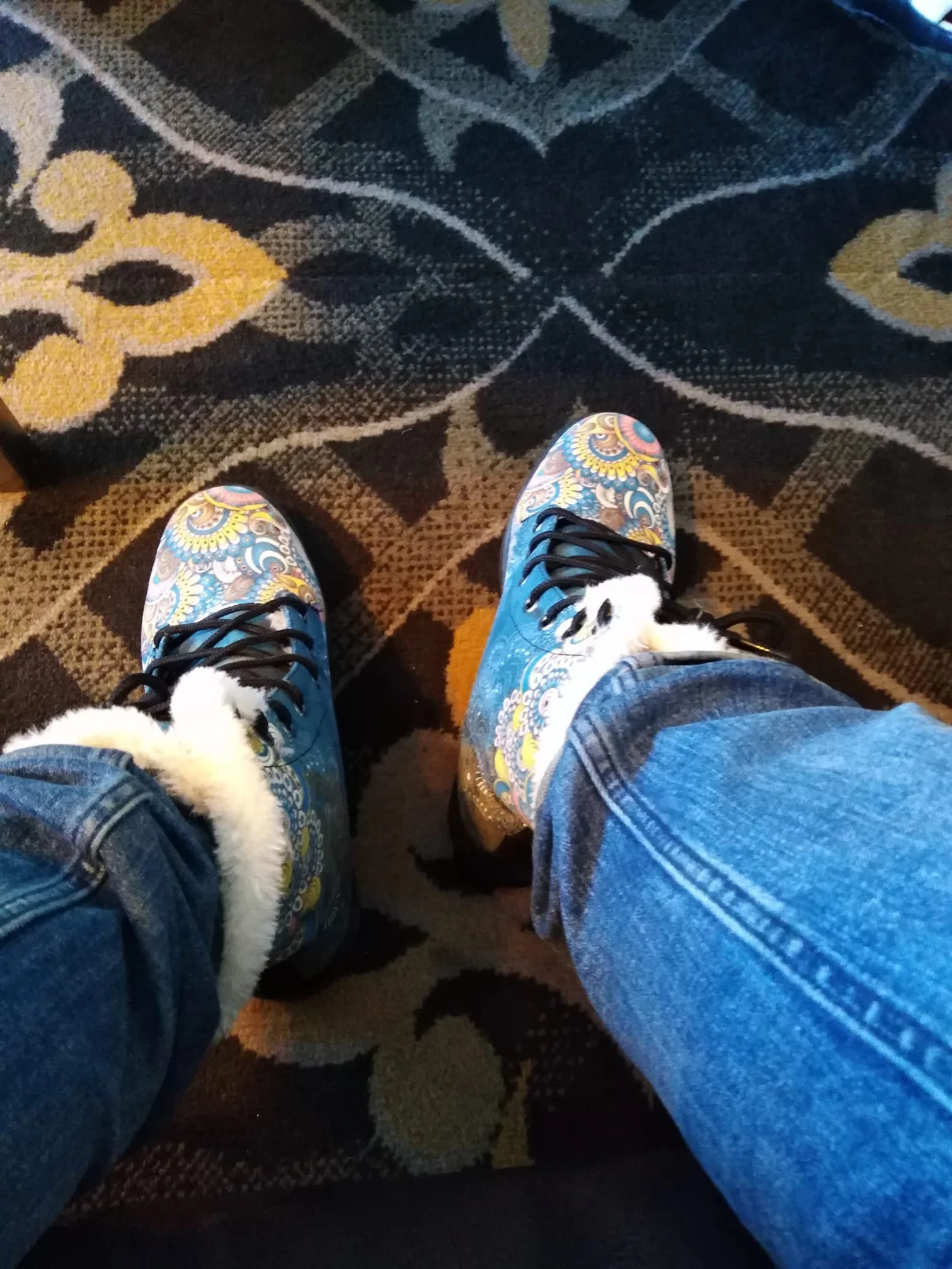 Person wearing colorful, patterned boots with faux fur trim, standing on a patterned carpet.