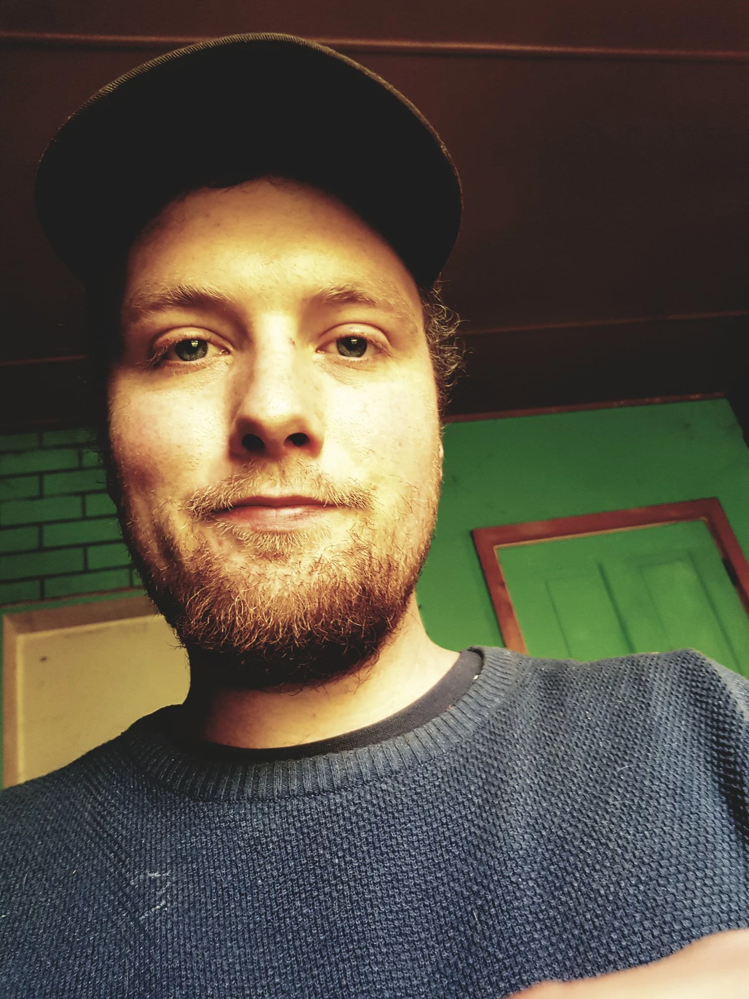 Close-up selfie of a young man with a beard, wearing a black baseball cap and a dark textured sweater, standing indoors with a green and red framed wall background.