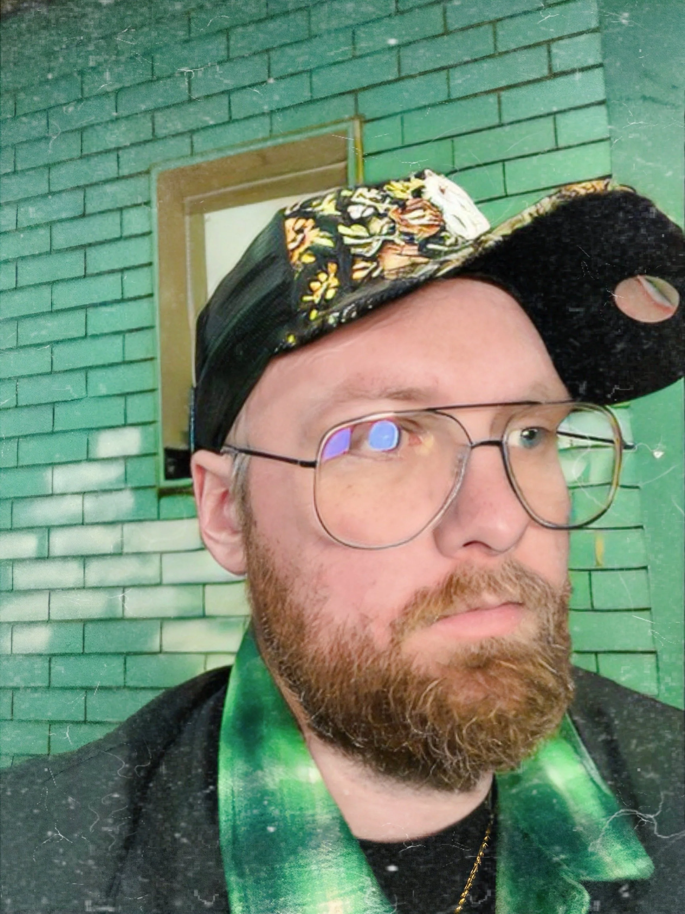 A man with glasses and a beard wearing a black cap with a floral pattern and a green scarf, standing in front of a green brick wall with a mirror behind him.