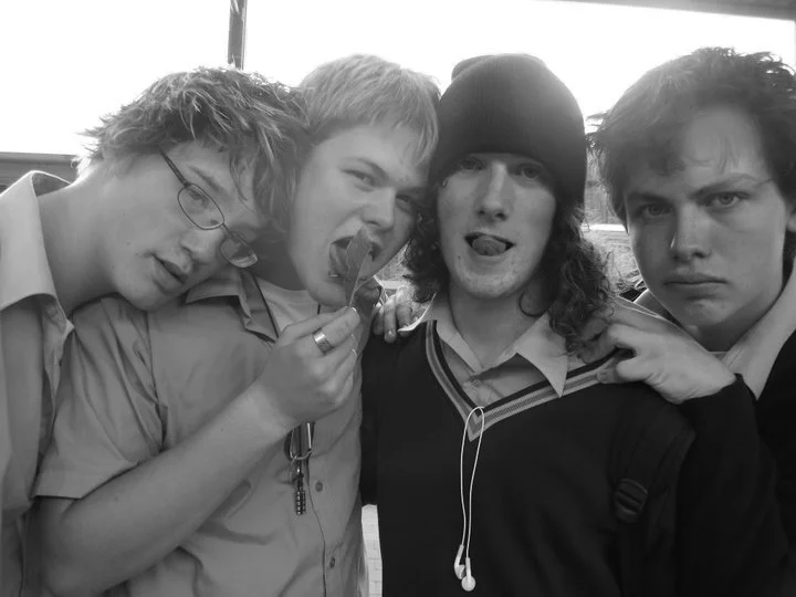 Four young men posing closely together, with three making serious or playful faces and one sticking out his tongue, in an indoor setting.