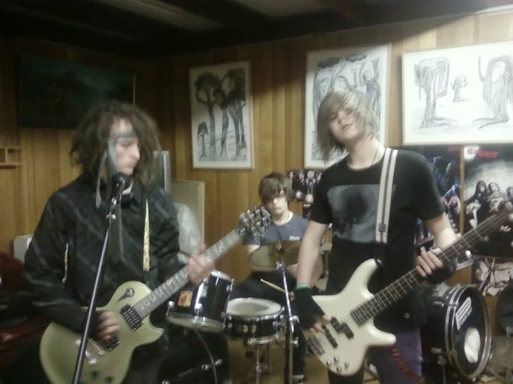 Three young musicians playing guitar and drums in a room with wood-paneled walls and framed artwork.