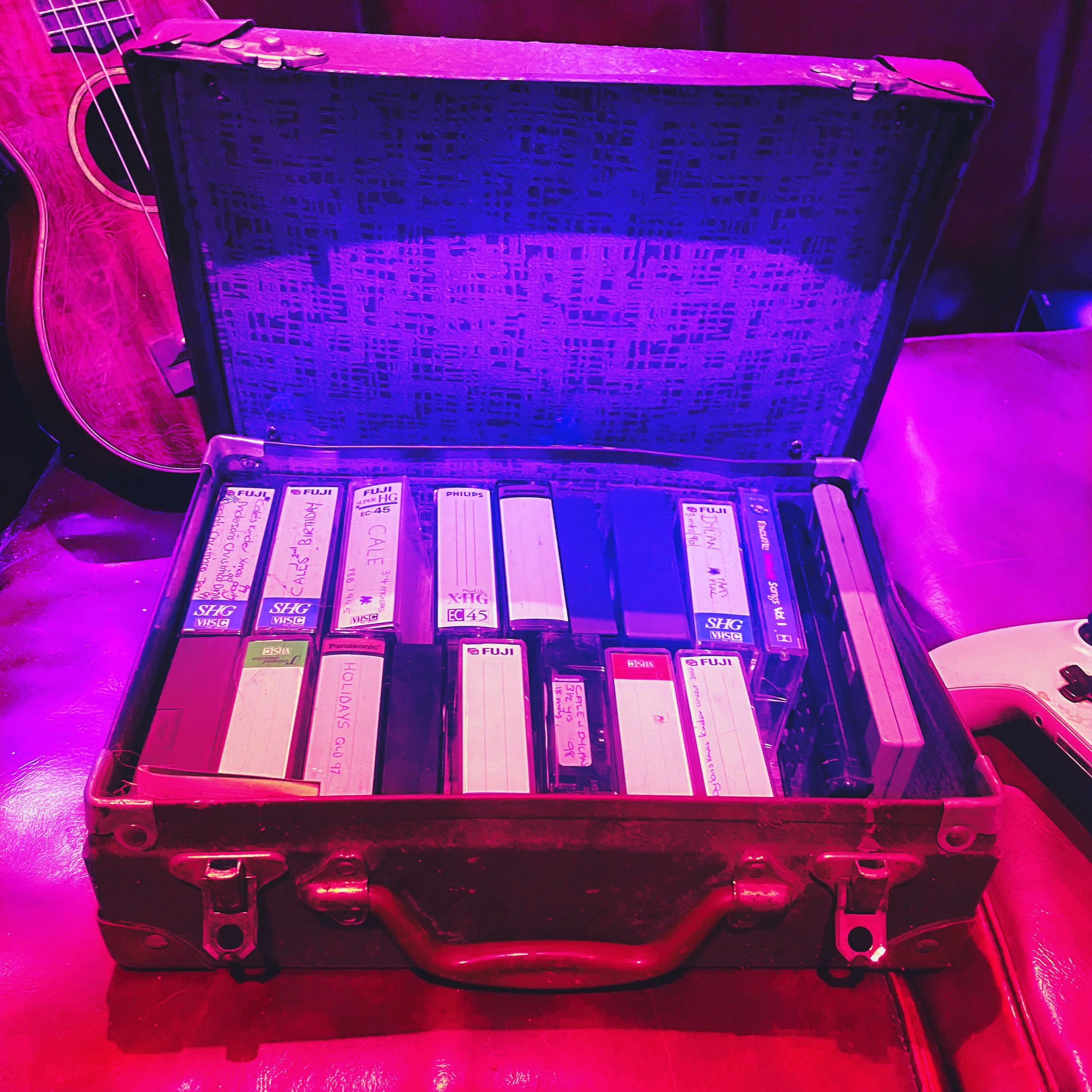 An open vintage metal briefcase containing several audio cassette tapes with handwritten labels, placed on a leather surface with a guitar in the background.