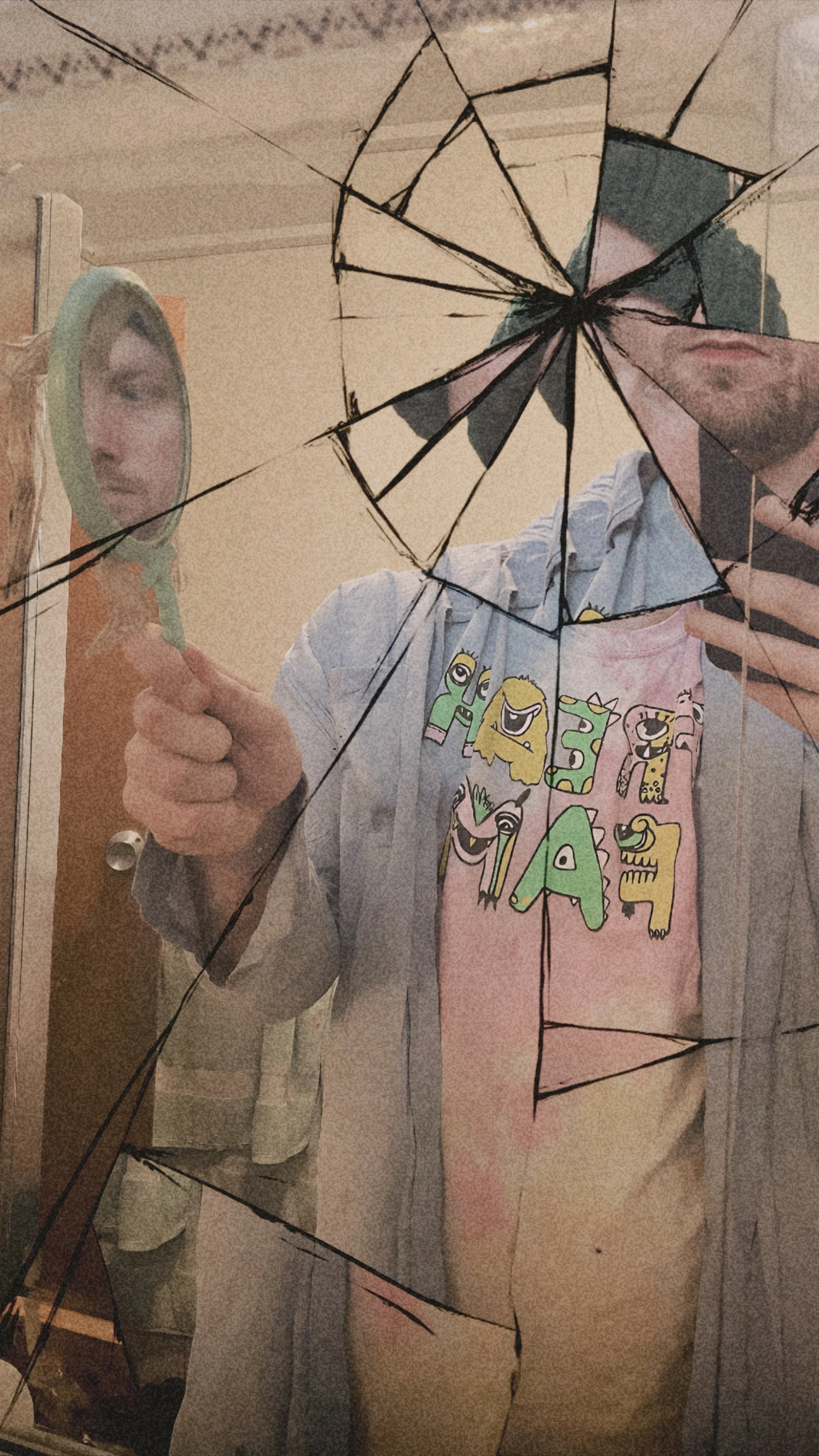 Person taking a selfie in a cracked mirror, holding a mirror with a magnifying side, wearing a colorful and playful T-shirt, with part of their face and hand visible.