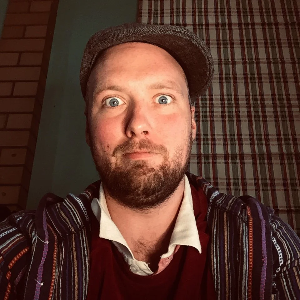 Close-up portrait of a man with blue eyes, light skin, a beard, and wearing a brown cap, a white shirt, and a patterned jacket, standing in front of a brick wall and a plaid curtain.