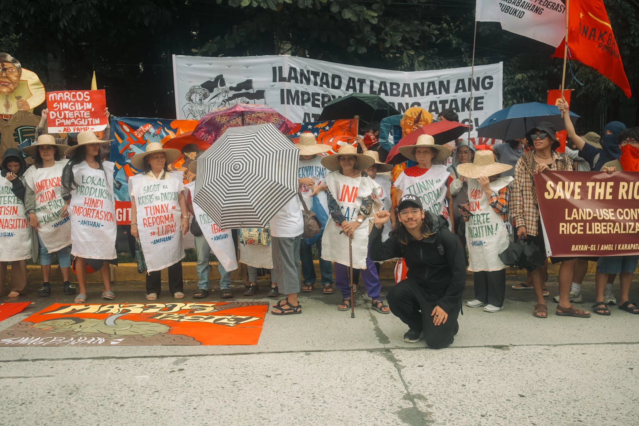 A group of people protesting with banners and signs. They are mostly women, some wearing straw hats, holding umbrellas, and dressed in white shirts with messages. Some signs are written in Filipino, addressing issues like land use, rice liberalization, and political concerns. A man in front is kneeling and making a fist, smiling at the camera. The protest is outdoors on a sidewalk with trees and banners in the background.