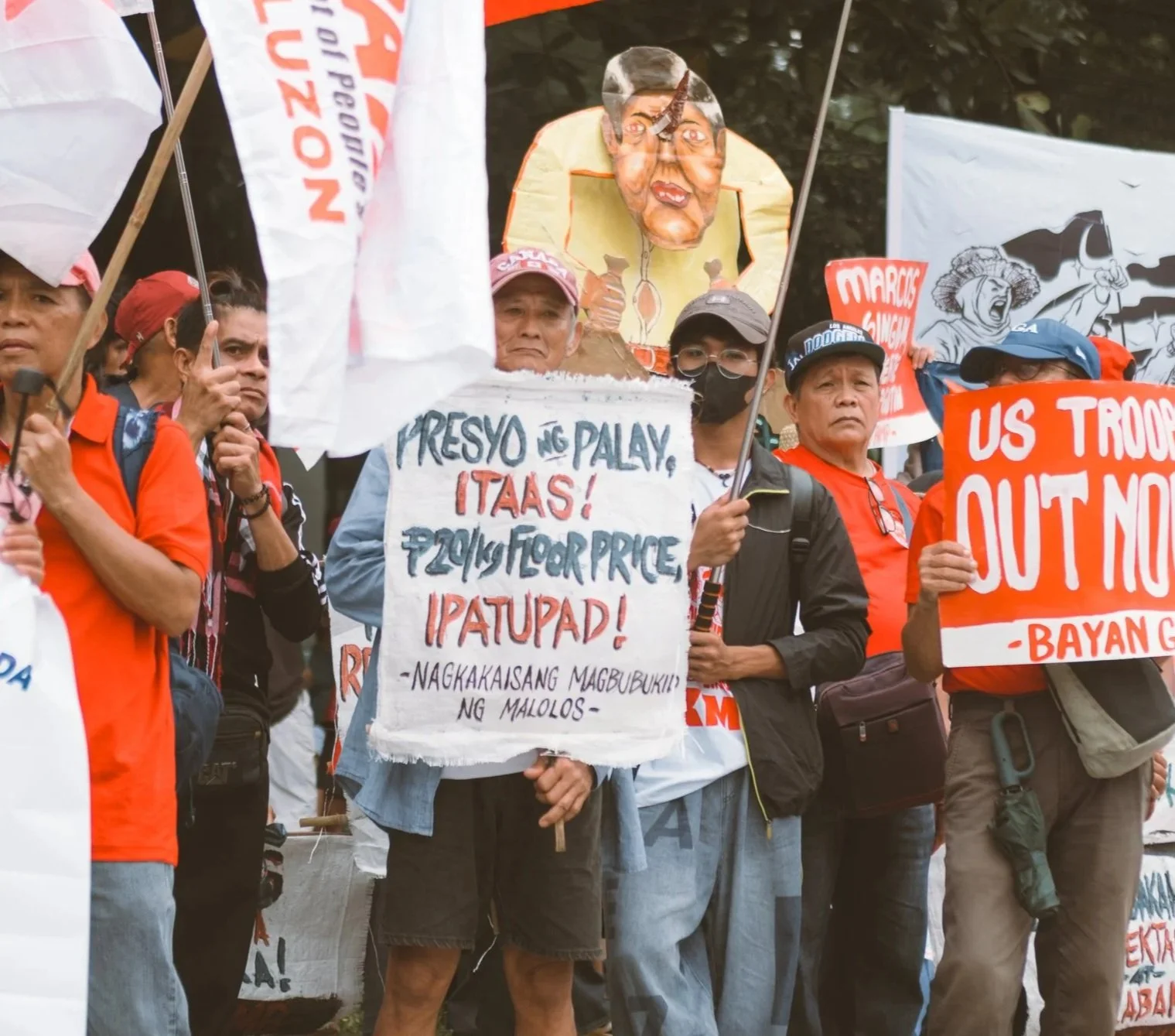 A group of protesters holding signs and banners during a demonstration. Some signs are written in Filipino, and there are masks and flags among them.