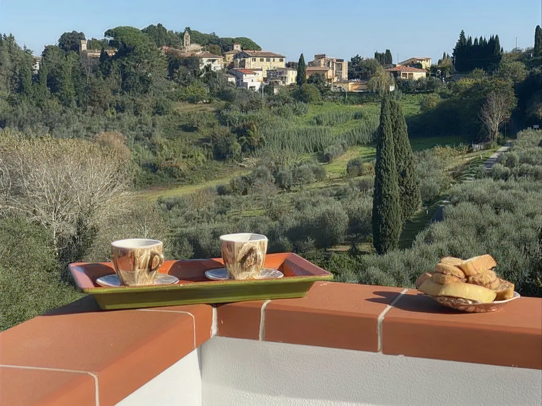 Two coffee cups on a tray with cookies on a balcony ledge overlooking a hilly landscape with trees, shrubs, and houses in the distance.