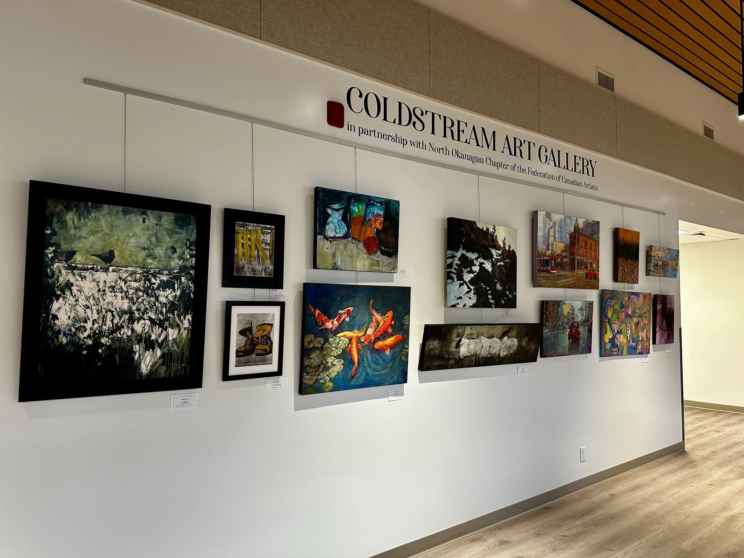 Display of various colorful artworks on a white gallery wall at Coldstream Art Gallery, with a sign above that reads 'Coldstream Art Gallery in partnership with North Okanagan Chapter of the Federation of Canadian Artists'.