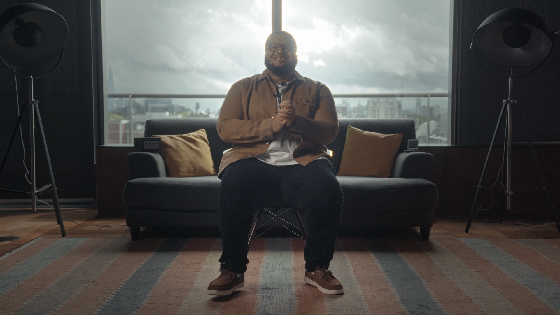 Goodness Akalazu, Founder of Immersely, A man sitting in front of a large window with a city view, smiling, wearing glasses, a tan jacket, dark pants, and sneakers, with two large studio lights on tripods on either side of him.