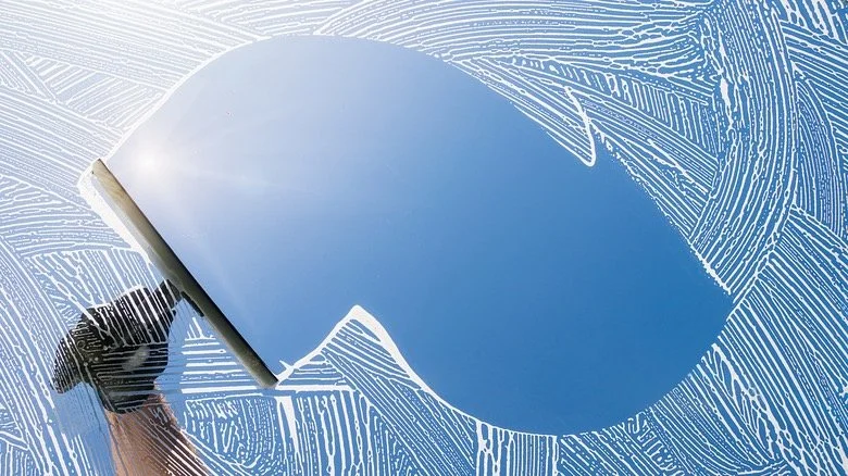 A person using a squeegee to perform exterior cleaning on a large glass window, creating a clear path through soap suds with sunlight reflecting off the glass.