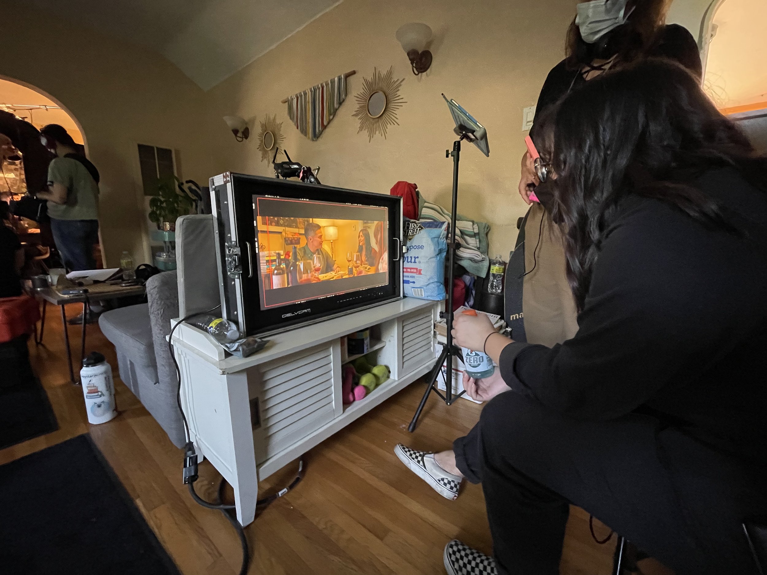 Group of people on a film set inside a house, with a monitor displaying a scene. One person is seated, holding a drink can, viewing the monitor. Equipment and bags are visible around the room.